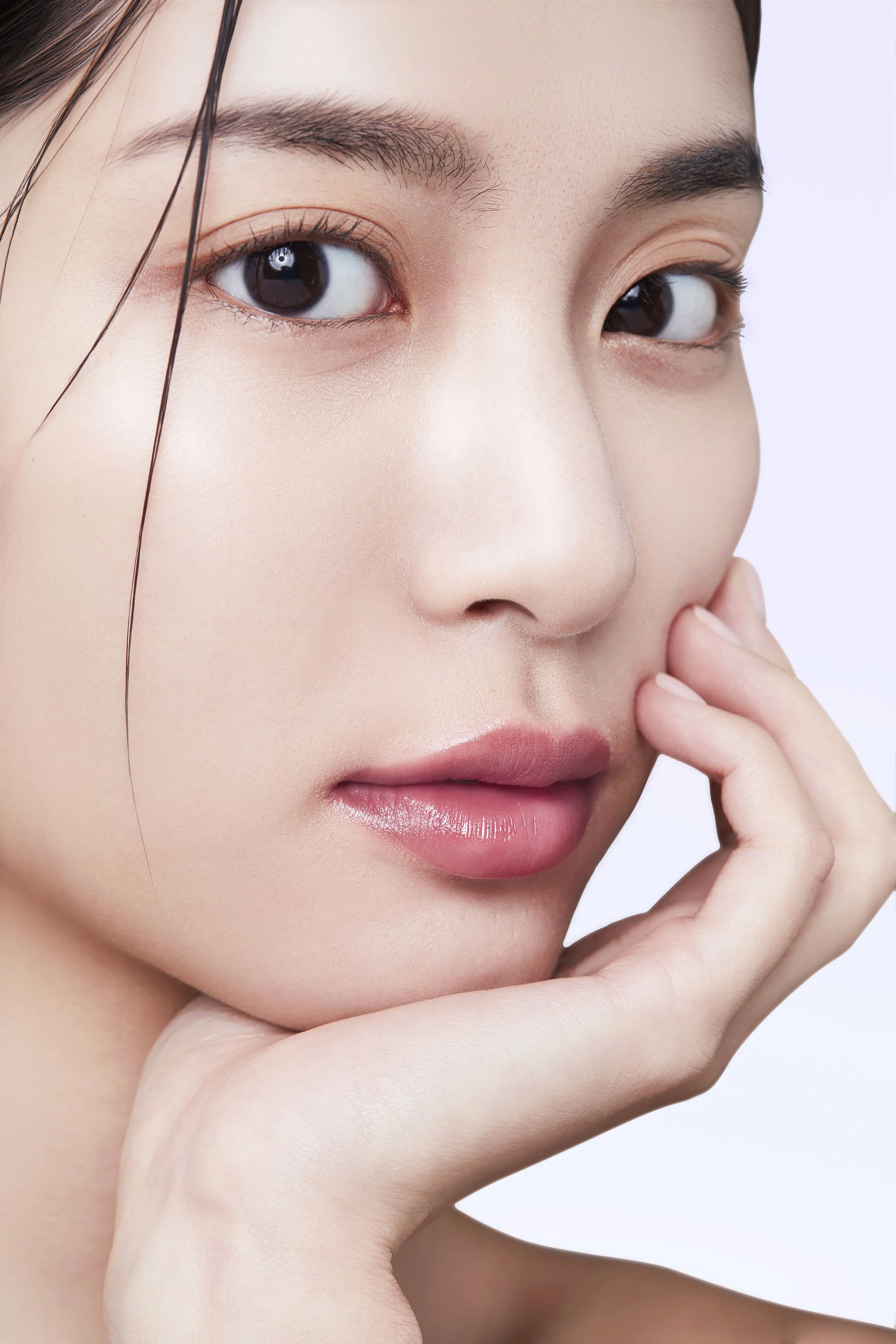 Close-up of a woman with flawless skin, dark eyes, and groomed eyebrows, touching her face gently.