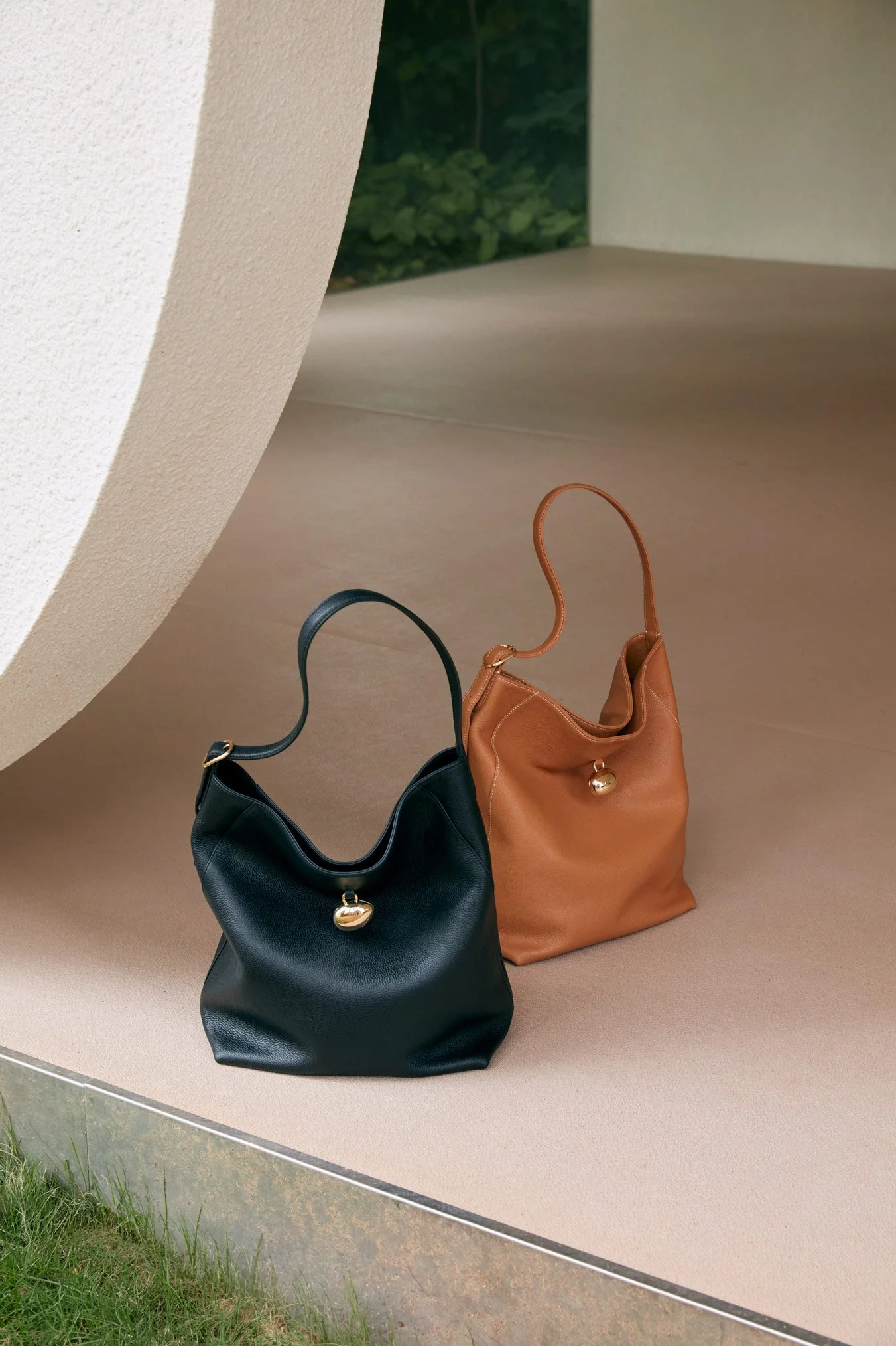 Two handbags, one black and one tan with a gold heart charm, placed on a beige display surface with a background of green foliage visible through a cutout.