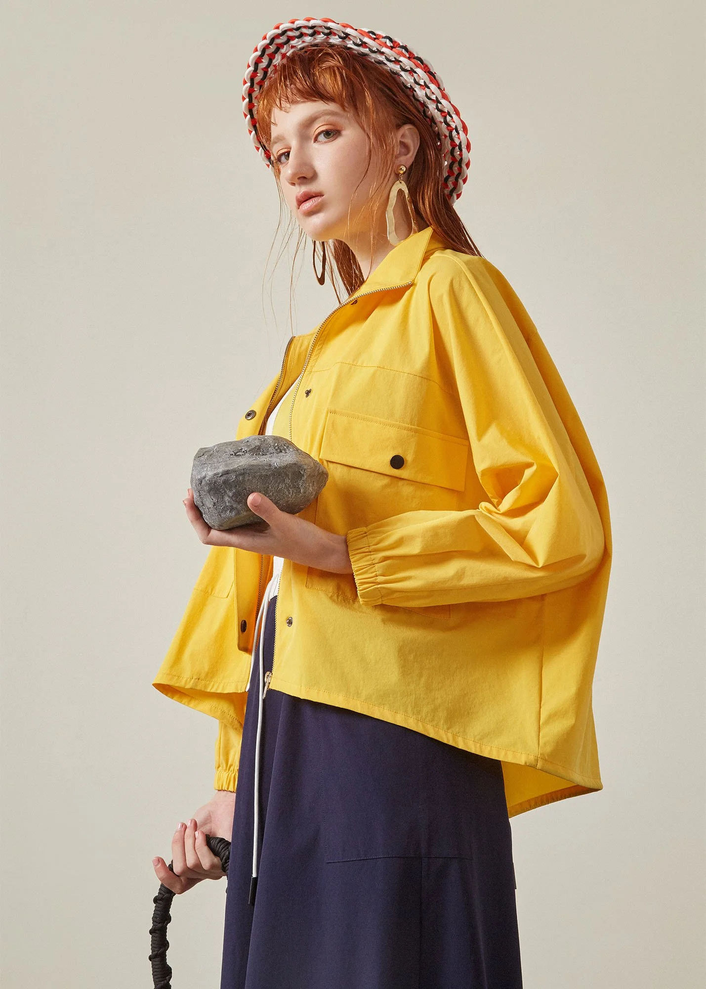 A young woman with red hair wearing a wide-brimmed hat with a red, white, and black pattern, gold earrings, a yellow jacket, and a navy skirt, holding a gray rock and a jump rope.
