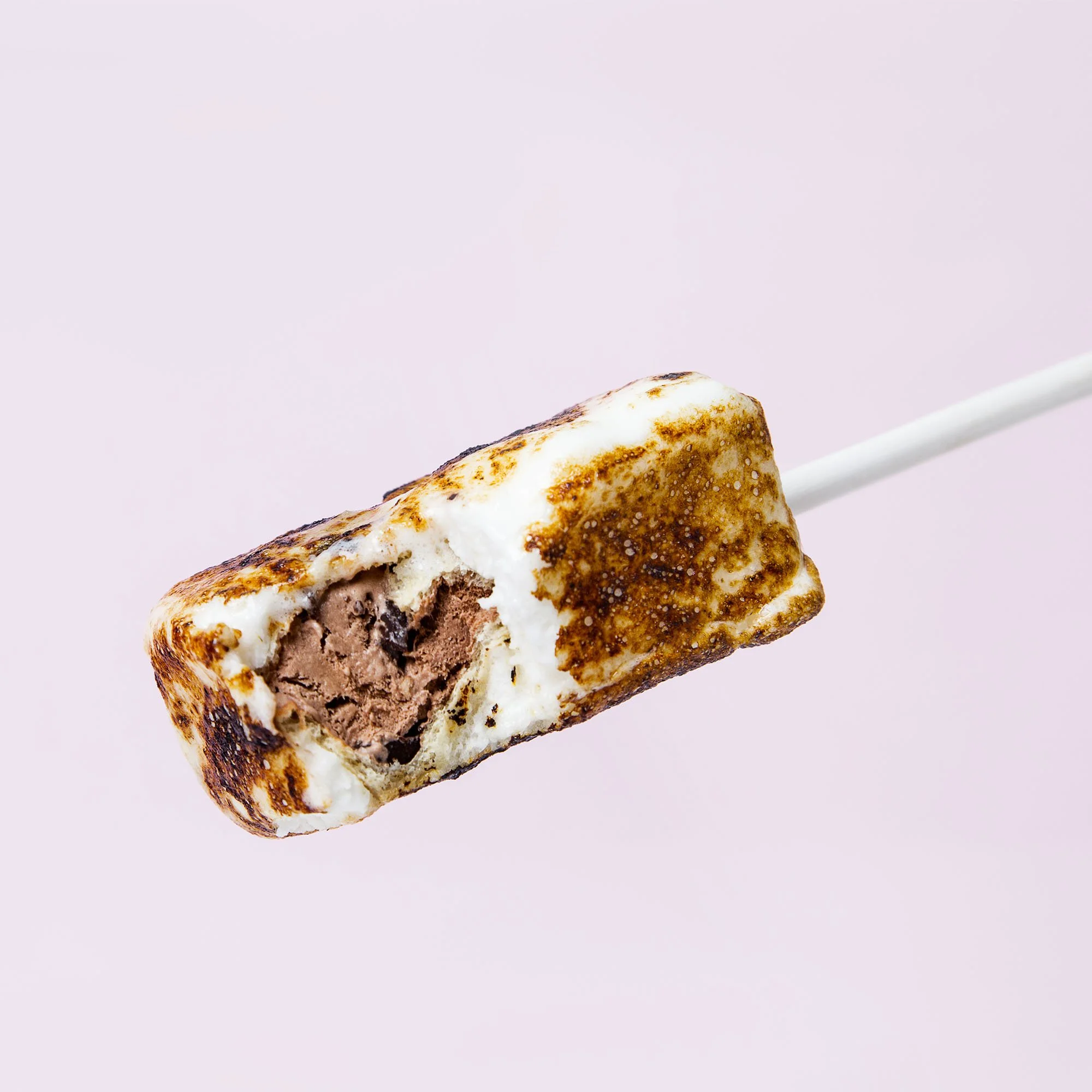 S'mores flavored ice cream bar with toasted marshmallow coating and chocolate ice cream inside on a stick against a pink background.