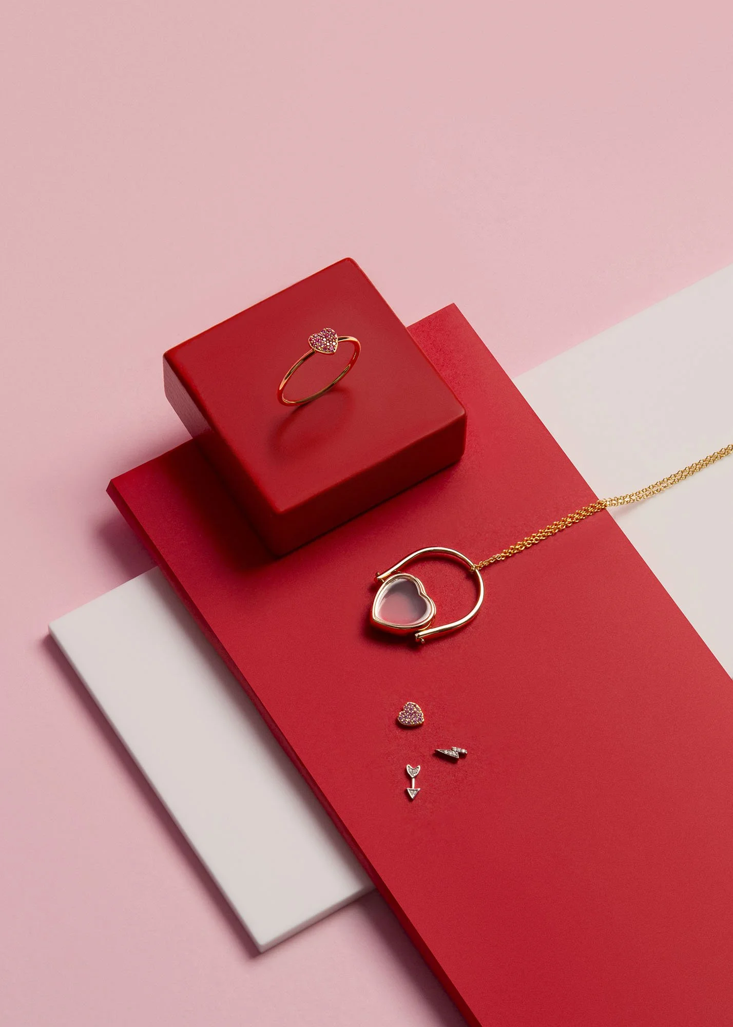 Gold jewelry set including a heart-shaped ring with pink stones, and a necklace with a gold heart-shaped pendant, displayed on red, pink, and white background boards with small decorative charms.