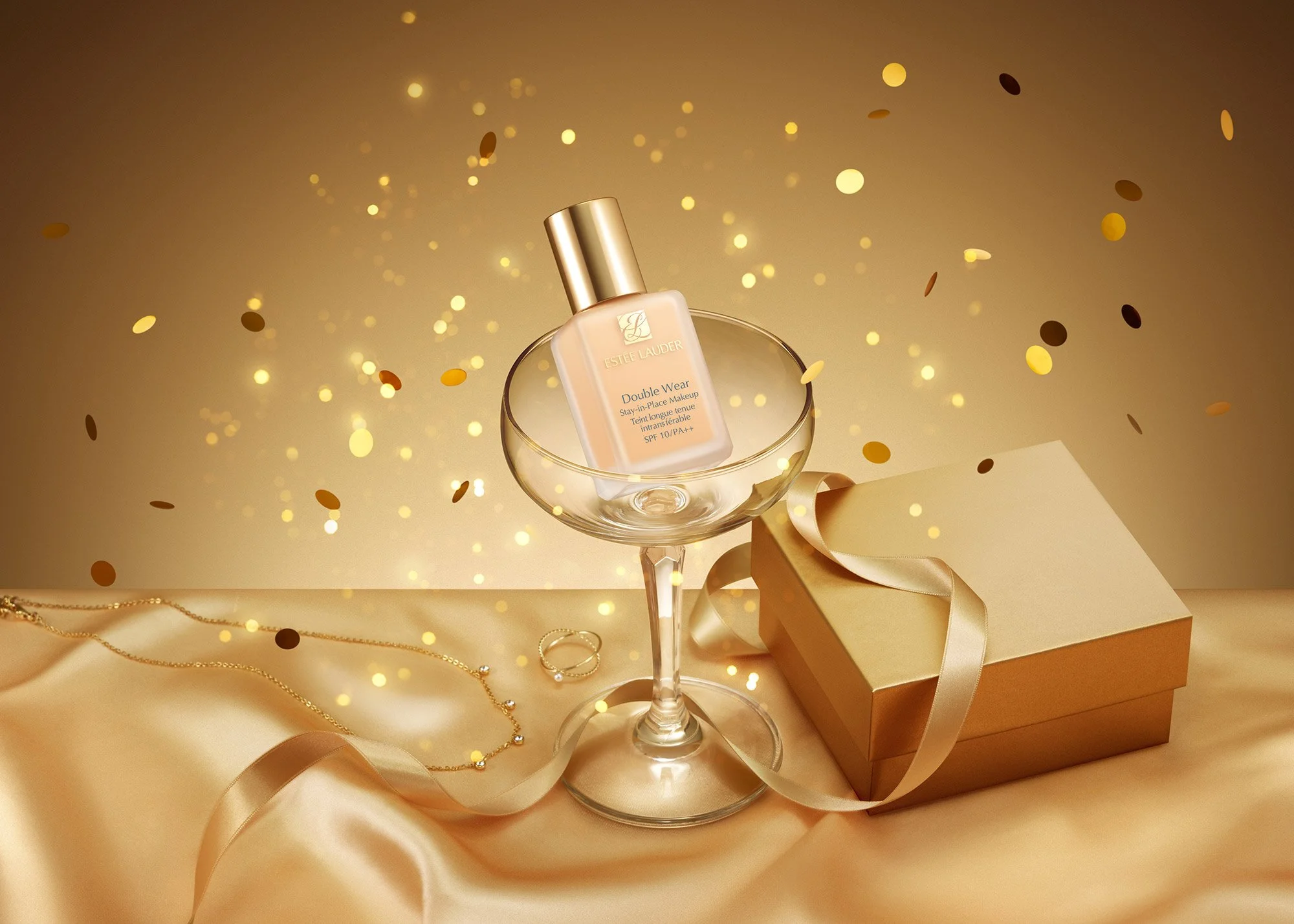 A bottle of Estee Lauder Double Wear foundation inside a cocktail glass with gold confetti falling around, placed next to a gold gift box with a satin ribbon, on a satin cloth, with jewelry in the background.