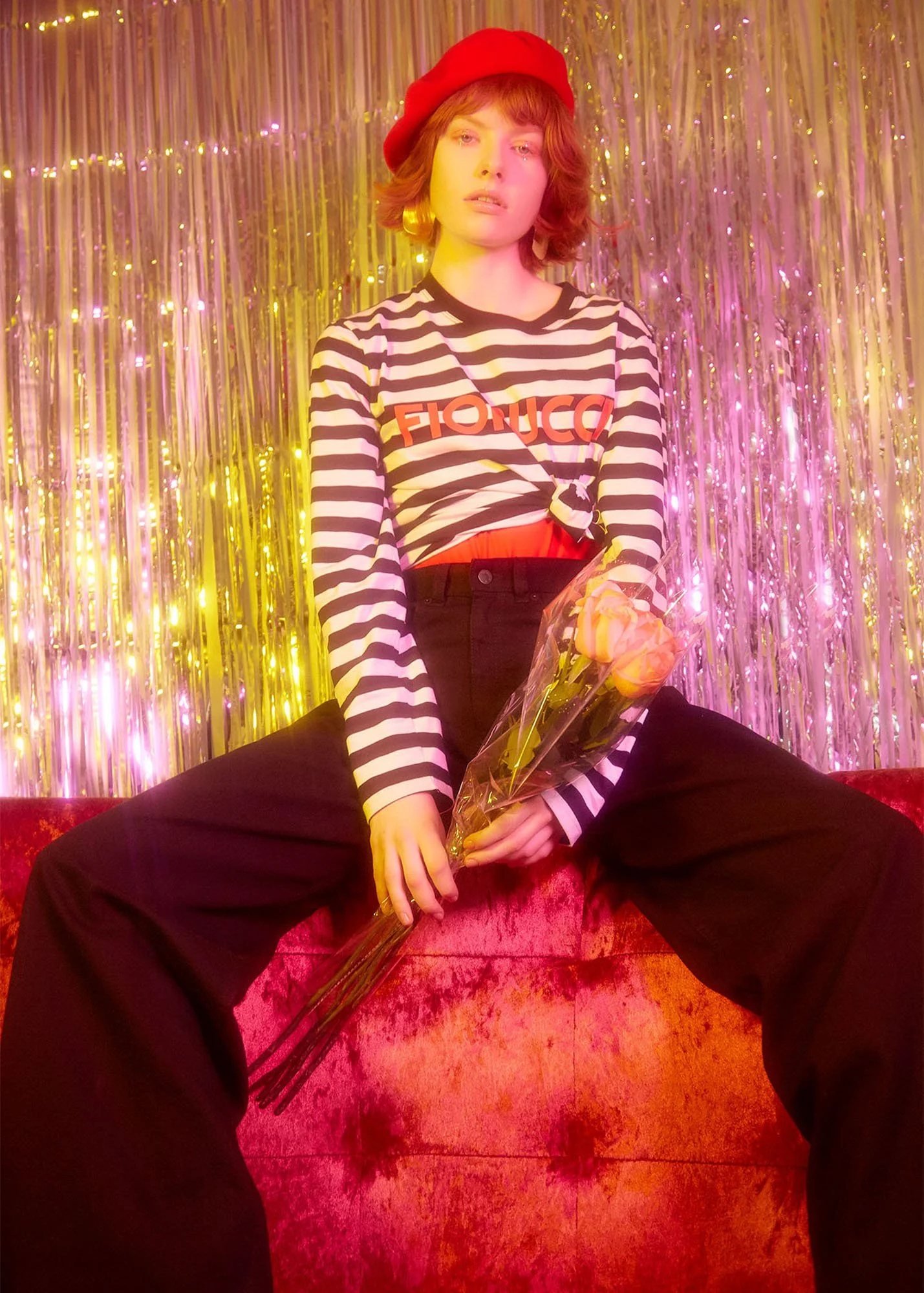 A woman with short wavy hair wearing a red beret, striped shirt with the word 'FIORE' on it, and black pants, sitting on a red velvet surface with legs spread apart. She is holding a bouquet of yellow roses wrapped in plastic. The background features