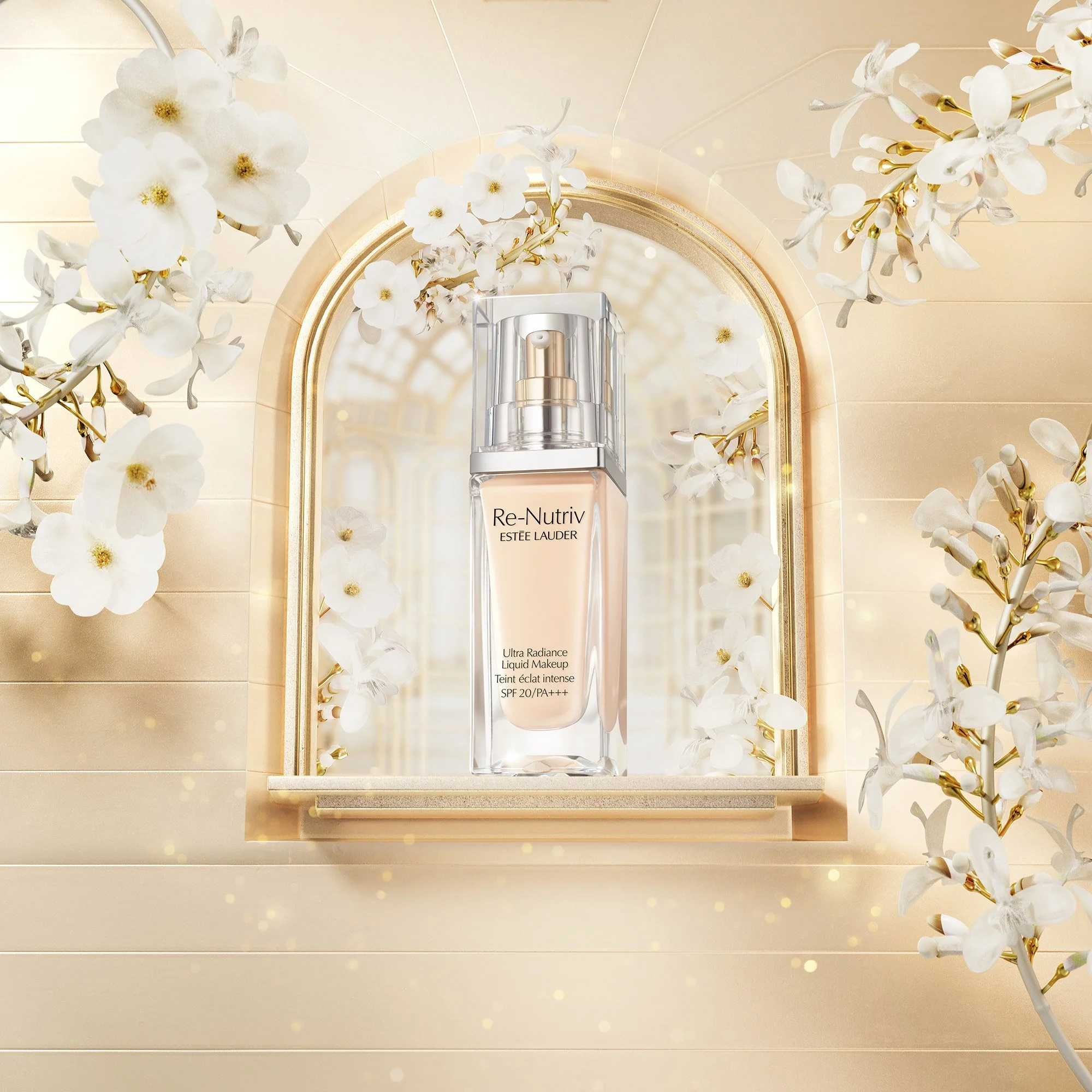 A bottle of Estée Lauder Re-Nutriv Ultra Radiance Liquid Makeup in front of a gilded arch mirror surrounded by white flowers with gold accents, set against a cream-colored wooden wall.