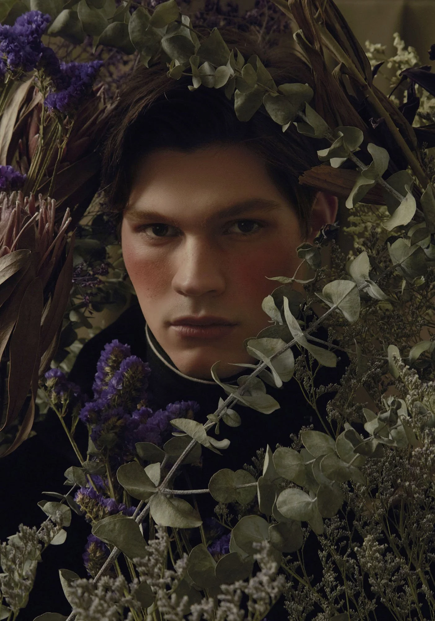 A young person with short dark hair surrounded by various flowers and green foliage, gazing intently at the camera.