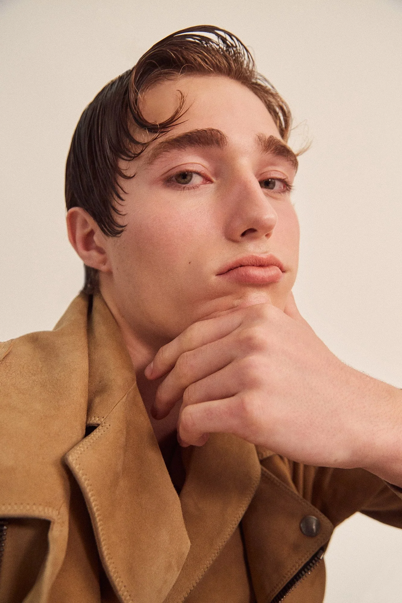 Close-up of a young man with slicked-back hair wearing a tan jacket, resting his chin on his hand, looking at the camera.