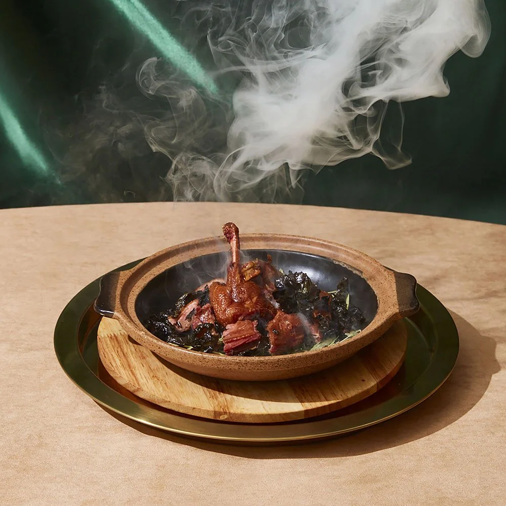 A sizzling dish with a cooked chicken leg and other meat pieces in a black skillet, placed on a wooden tray with steam and smoke rising from it.