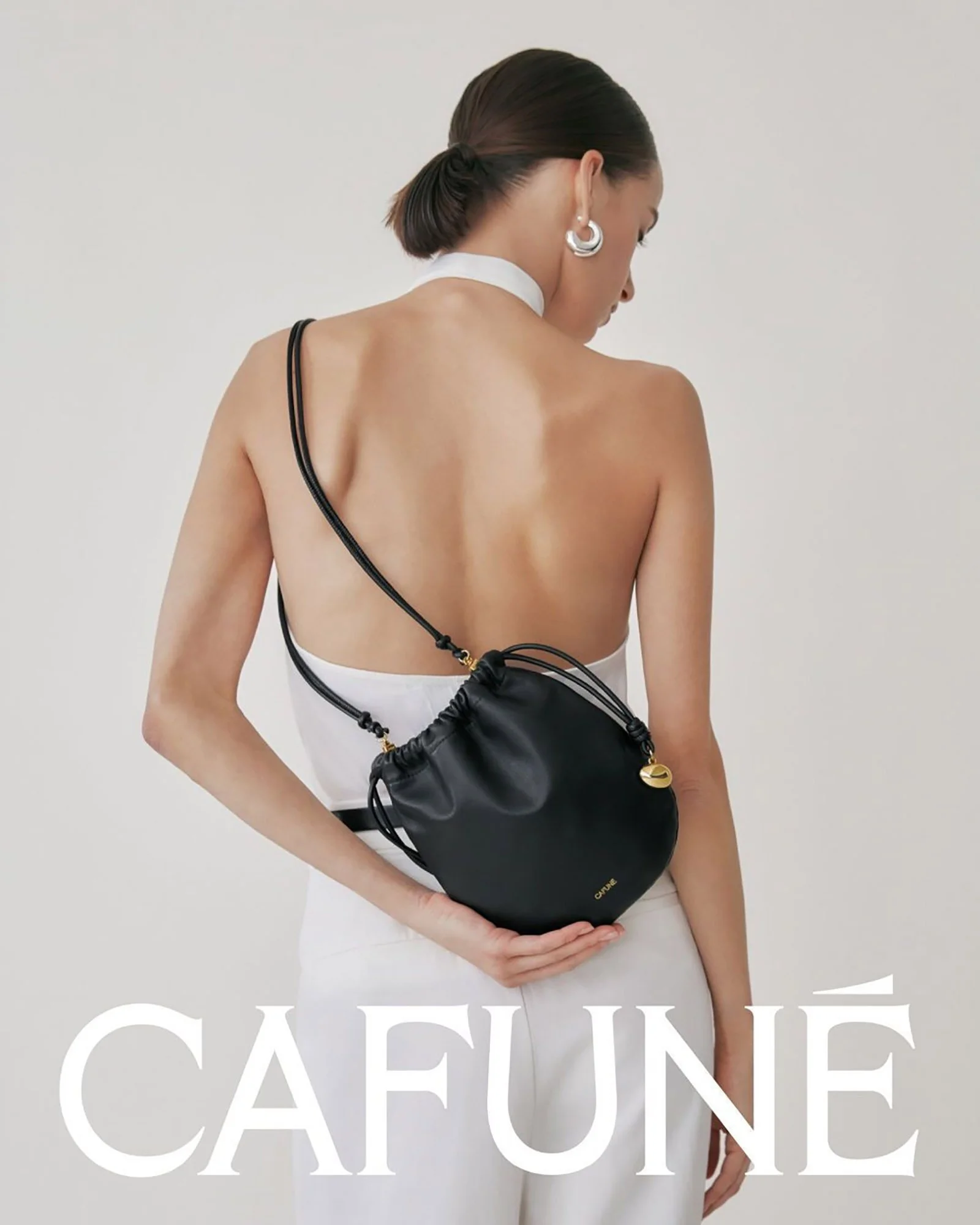 A woman with short brown hair styled in a low bun is wearing large silver hoop earrings and a white choker necklace. She is viewed from the back, with bare shoulders, and has a black drawstring bag hanging across her left shoulder. She is standing ag