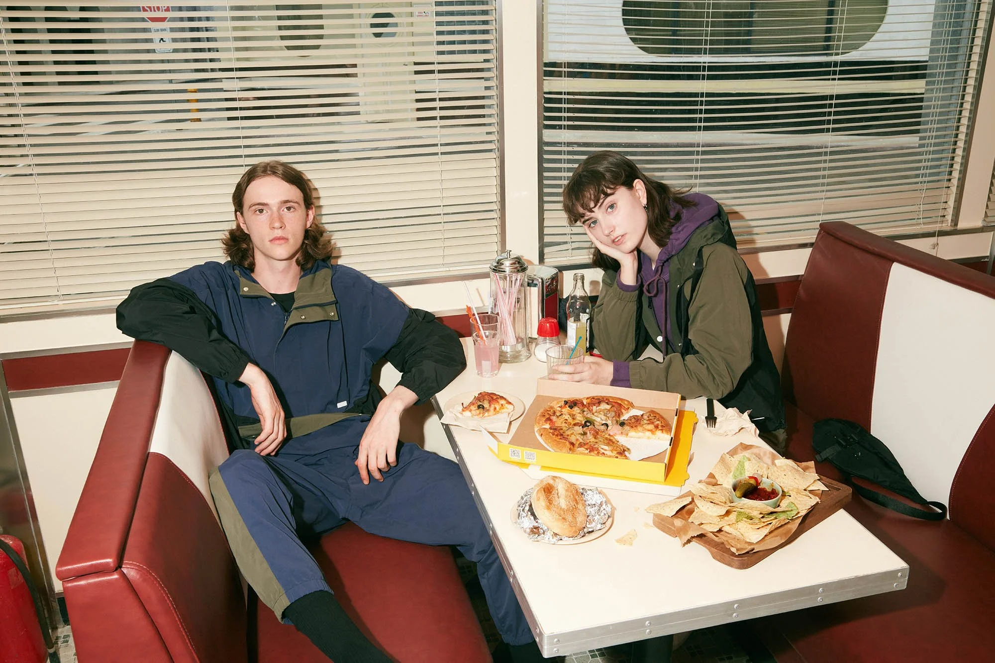 Two young people sitting in a booth at a pizza restaurant, with pizza, bread, and drinks on the table.