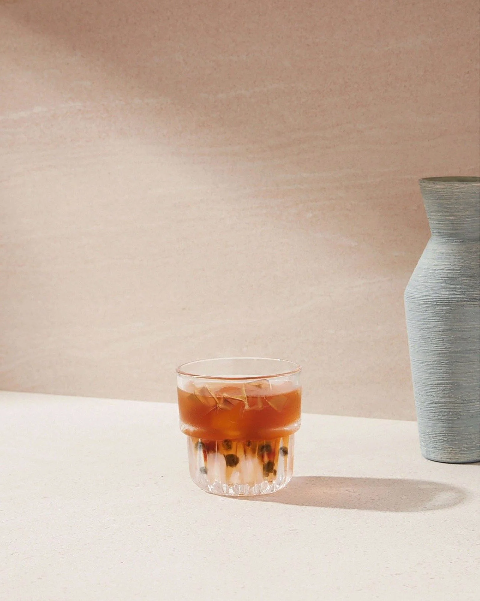 A glass of iced tea with bubbles and ice cubes on a white surface, with a tall grey textured vase to the right and a beige background.