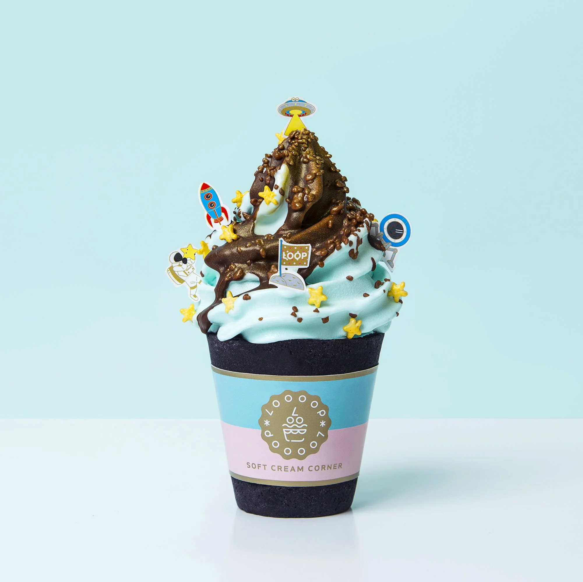 Ice cream cone with blue and pink wrapper, topped with chocolate and caramel sauces, sprinkles, and space-themed sprinkles like rockets, stars, and aliens, on a light blue background.