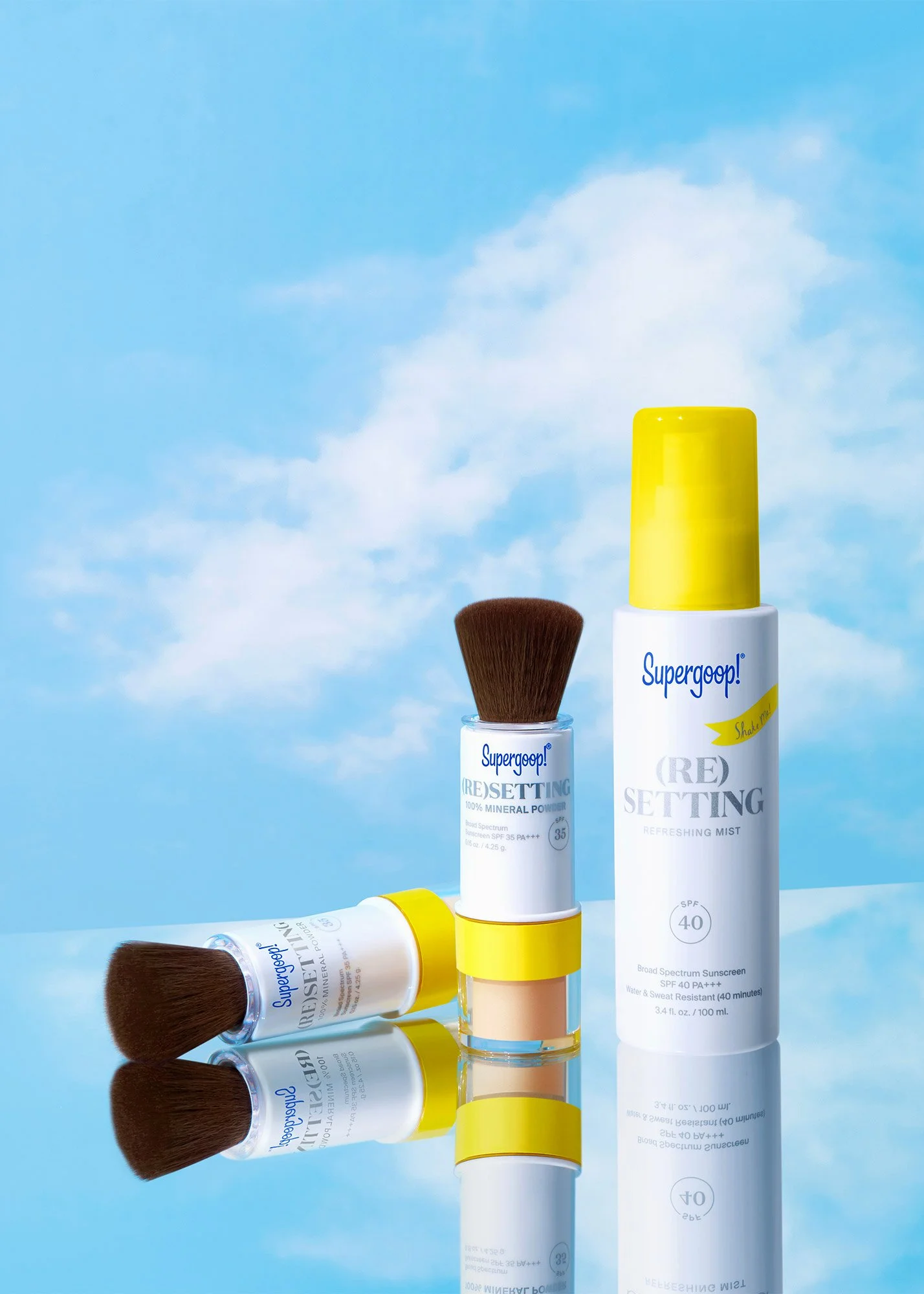 Three Supergoop! skincare products include two UV setting mists and one mineral powder, set against a background with a blue sky and clouds, with their reflections on a glossy surface.