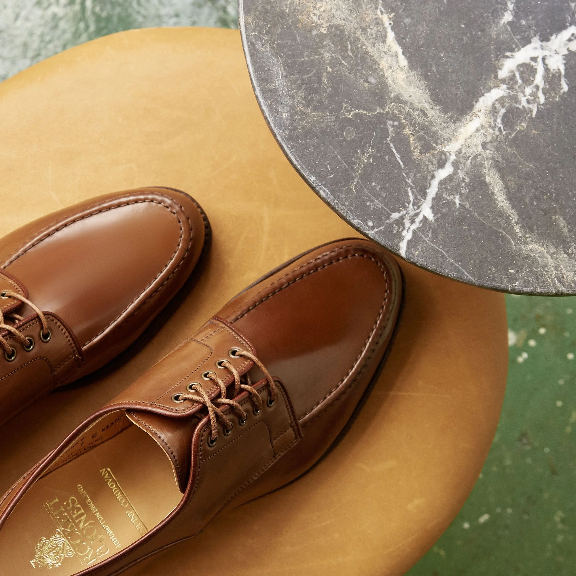 A pair of polished brown leather dress shoes placed on a brown leather surface with a black and white marble table partway in the frame.