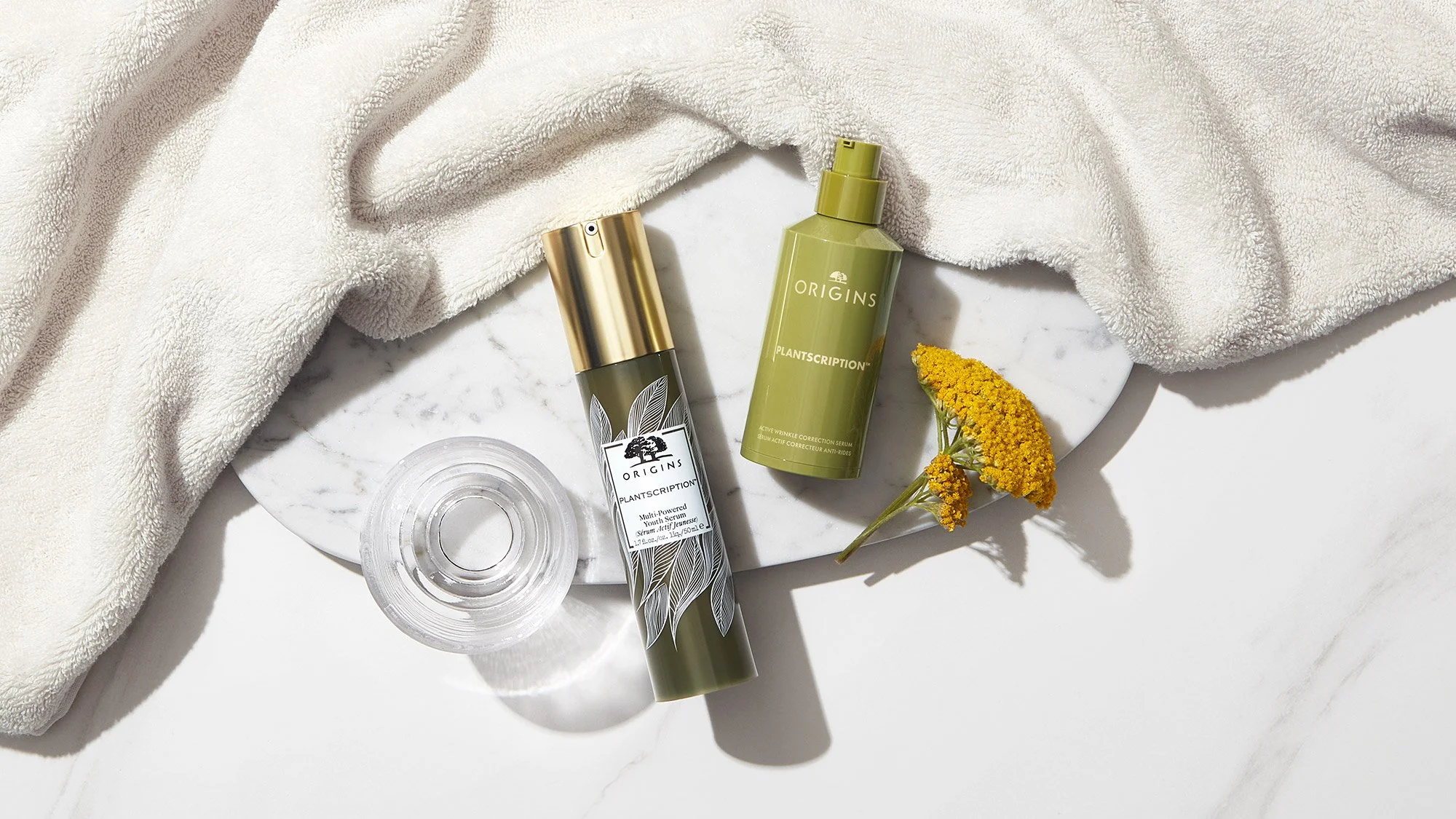 Skincare products from Origins Plantscription line, a clear round container, a small yellow flower, a white towel, and a white marble surface, with natural lighting.