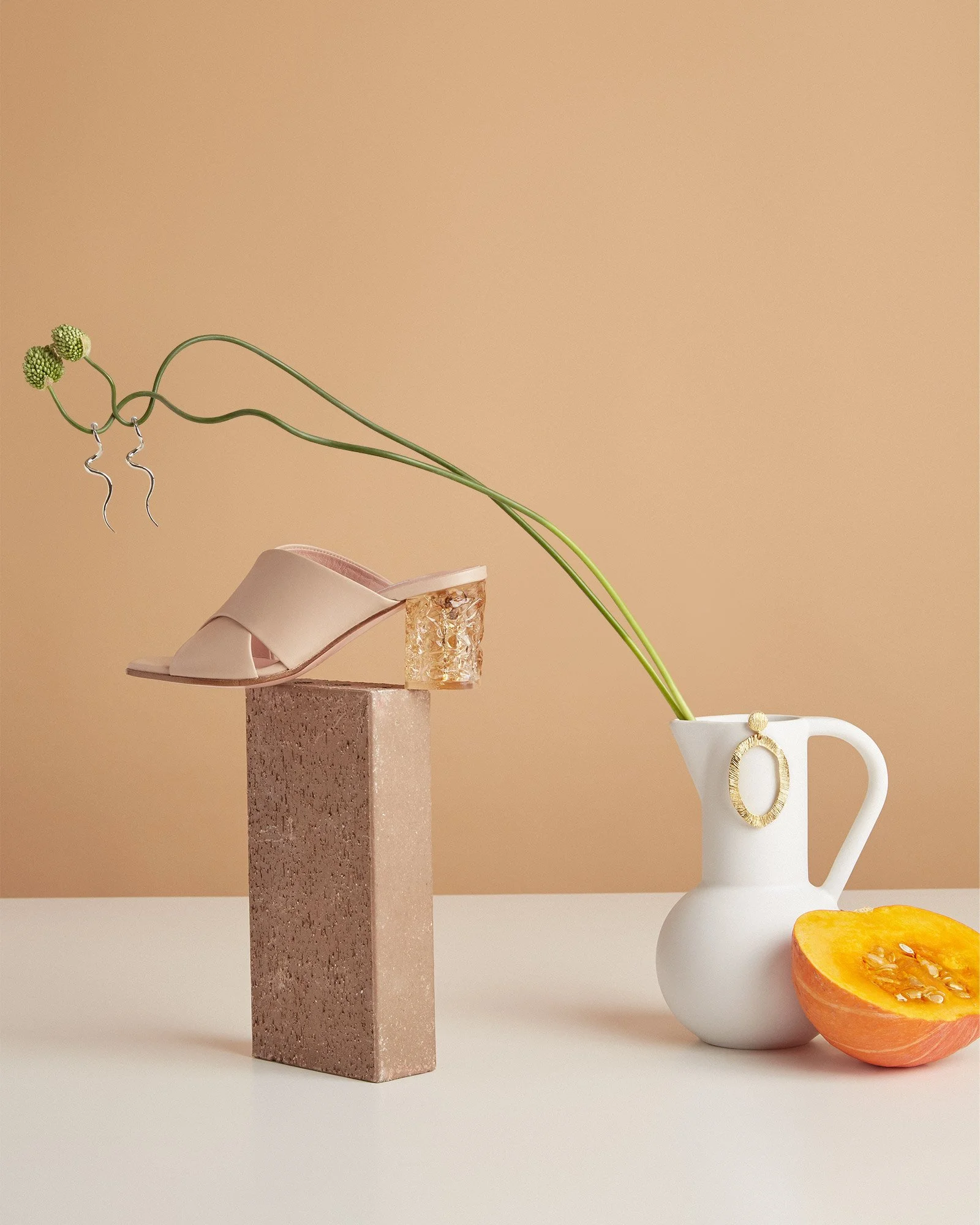 A still life composition featuring a white pitcher with a gold ring handle, a half-cut yellow pumpkin, a pink high-heeled sandal with a gold block heel, and a long green plant with small green flowers, all arranged on a neutral background.