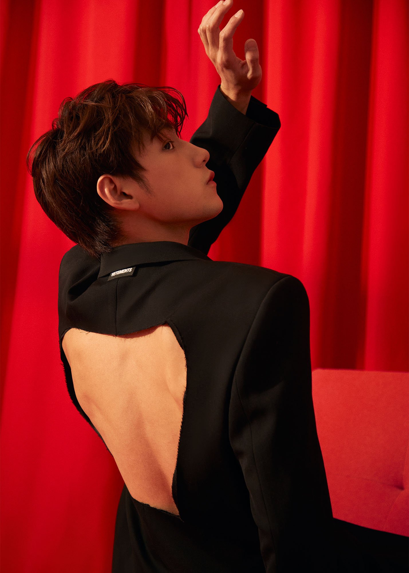 A person with short, tousled brown hair, wearing an asymmetrical black blazer with cutouts on the side, stands against a red curtain background, striking a pose with one arm raised and fingers elegantly poised.