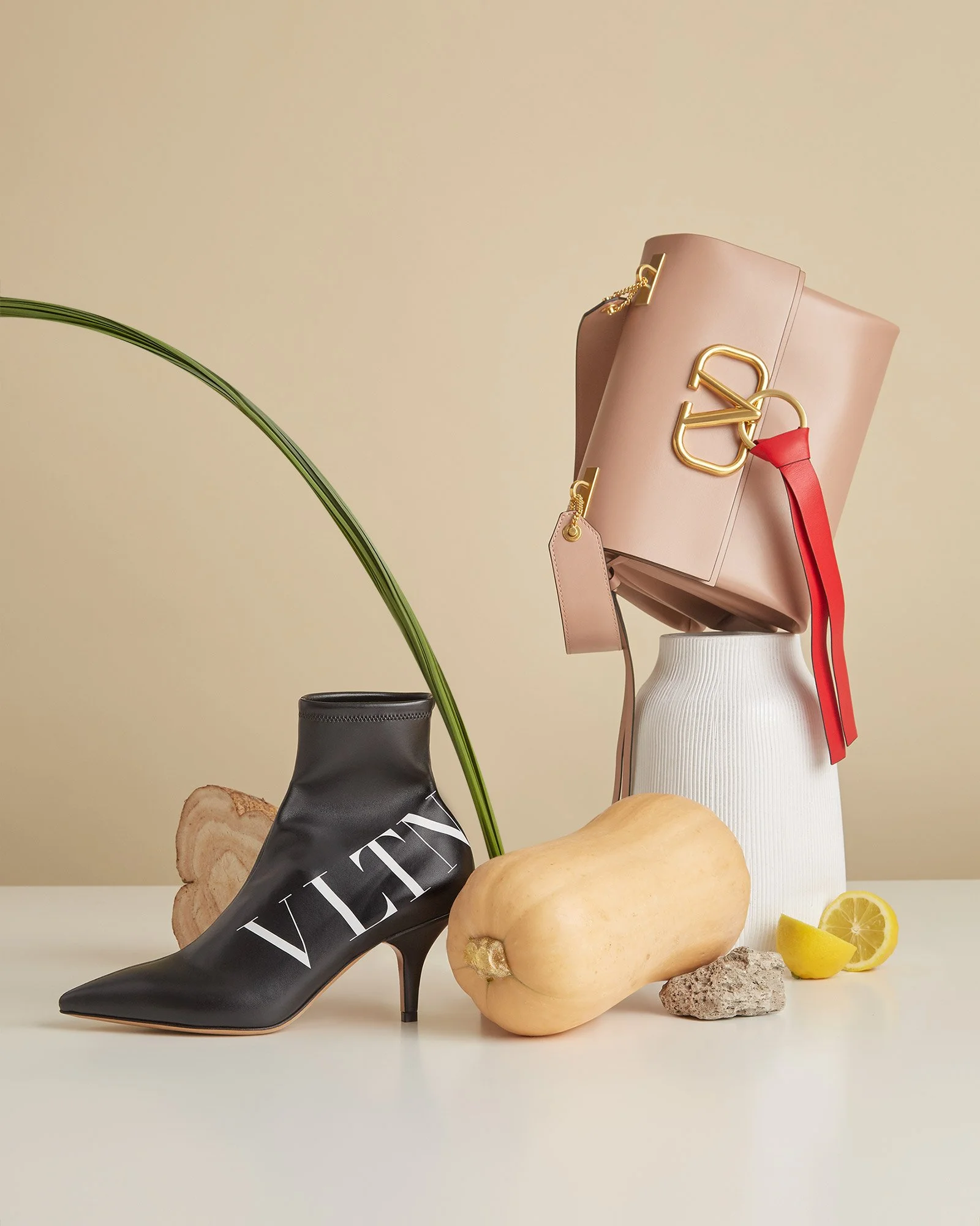 Fashion display featuring a black high-heeled ankle boot with white lettering, a beige pumpkin, a lime wedge, a rock, a tall white textured vase, a pink handbag with gold hardware and red ribbon, a beige tag, and a long green leaf, set against a plai