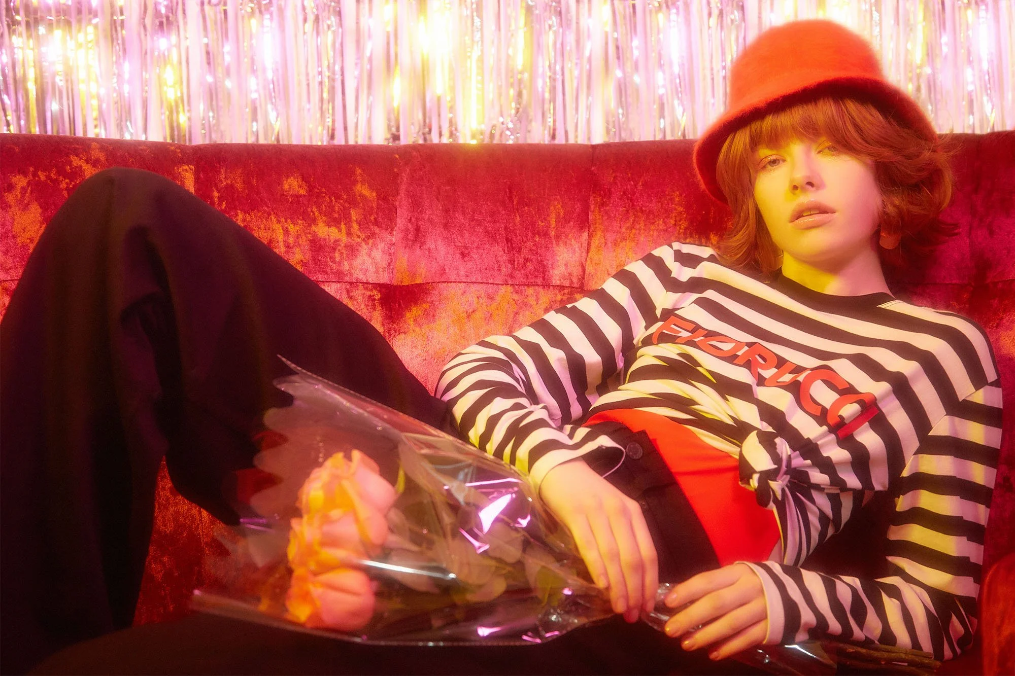 Woman lying on a red velvet couch holding a bouquet of pink roses, wearing a striped shirt and a red hat with colorful background.