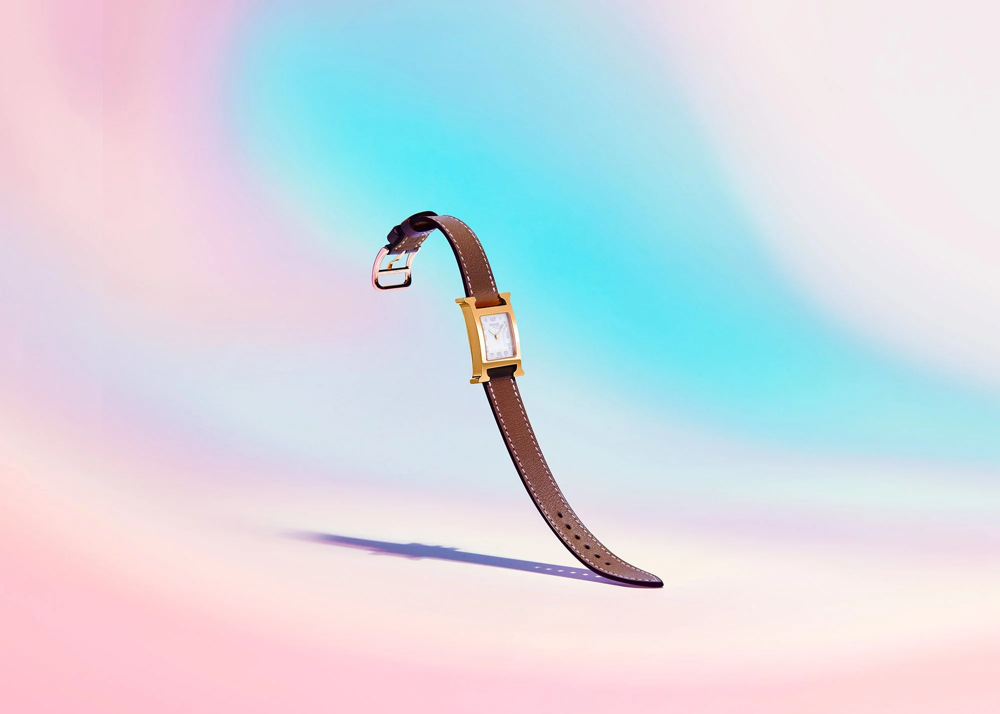 A wristwatch with a gold rectangular face and a brown leather strap, positioned at an angle against a pastel gradient background.