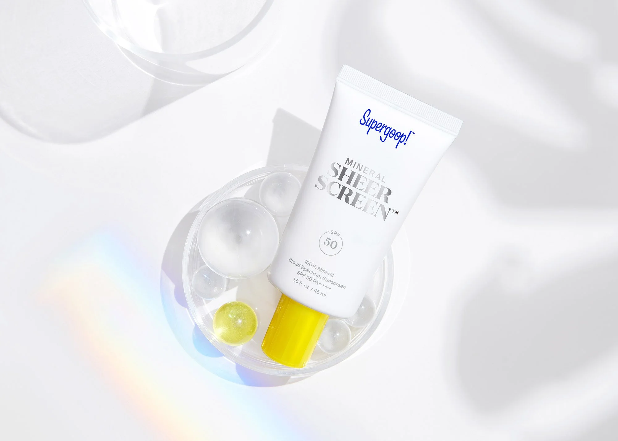 Tube of Supergoop! Mineral Sheer Screen SPF 50 sunscreen on a white surface with glass beads and a rainbow reflection.