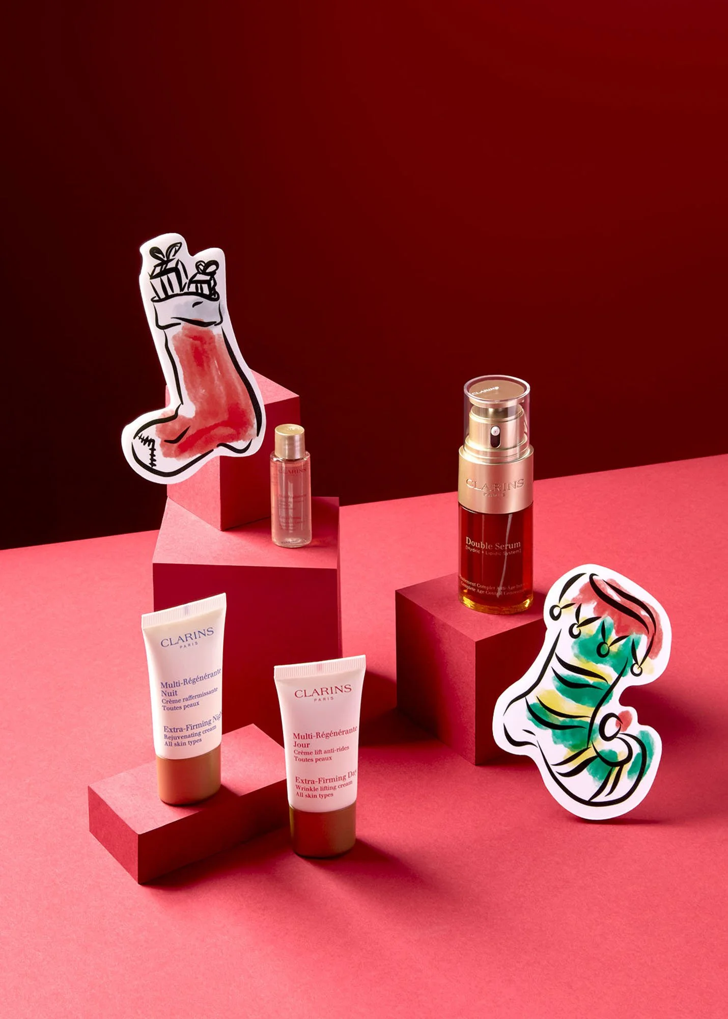 Arrangement of Clarins skincare products on pink platforms with two colorful seasonal-themed stickers in the background, against a red backdrop.
