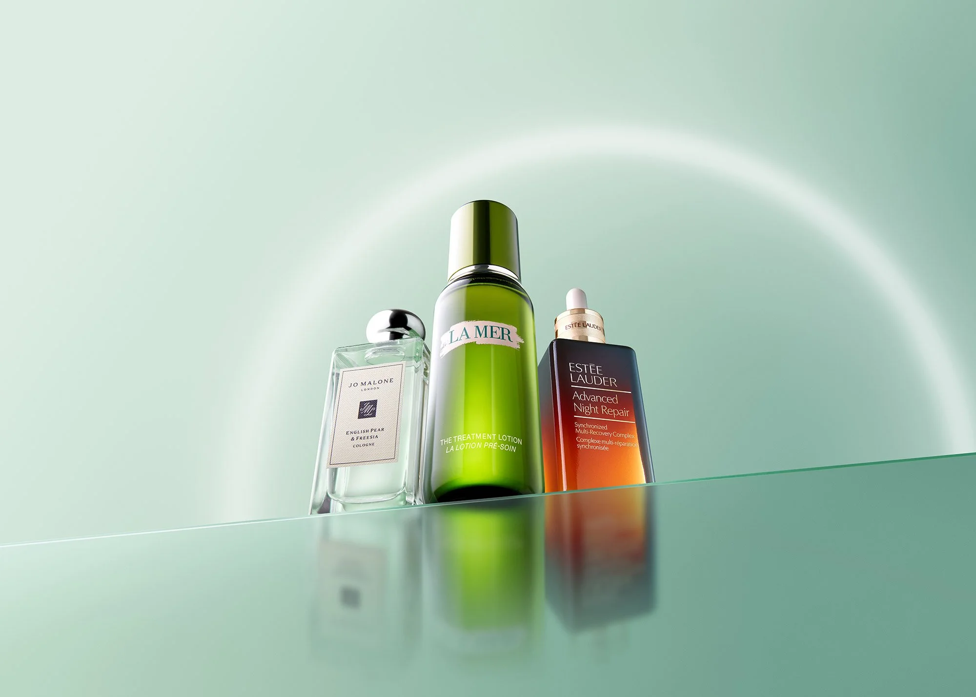 Three skincare products displayed on a reflective surface against a light green background with a subtle rainbow-like arc. The products include a Jo Malone cologne, a La Mer treatment lotion, and an Estée Lauder night repair serum.
