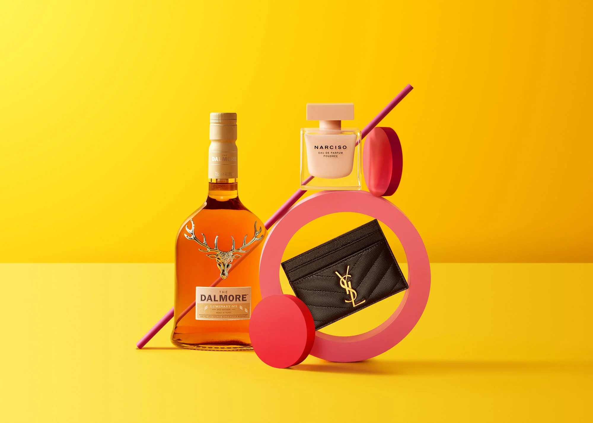 Arrangement of luxury items on a yellow background, including a bottle of Dalmore whiskey, a pink Narciso perfume, a black Yves Saint Laurent wallet, and geometric pink and red shapes.
