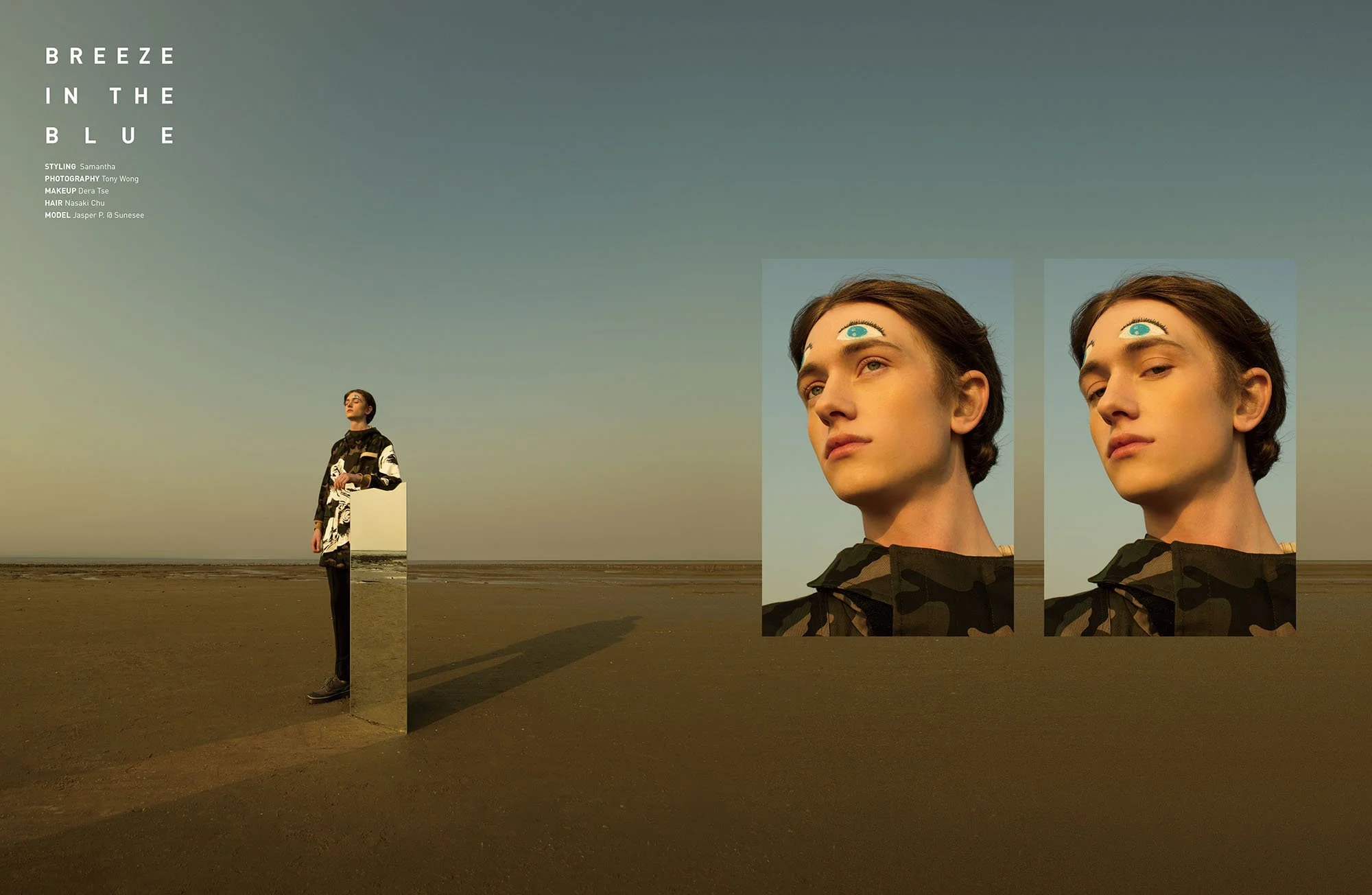 A person standing on a beach near a mirror and two digital screens displaying a close-up of the person's face with a third eye drawn on the forehead, against a twilight sky.