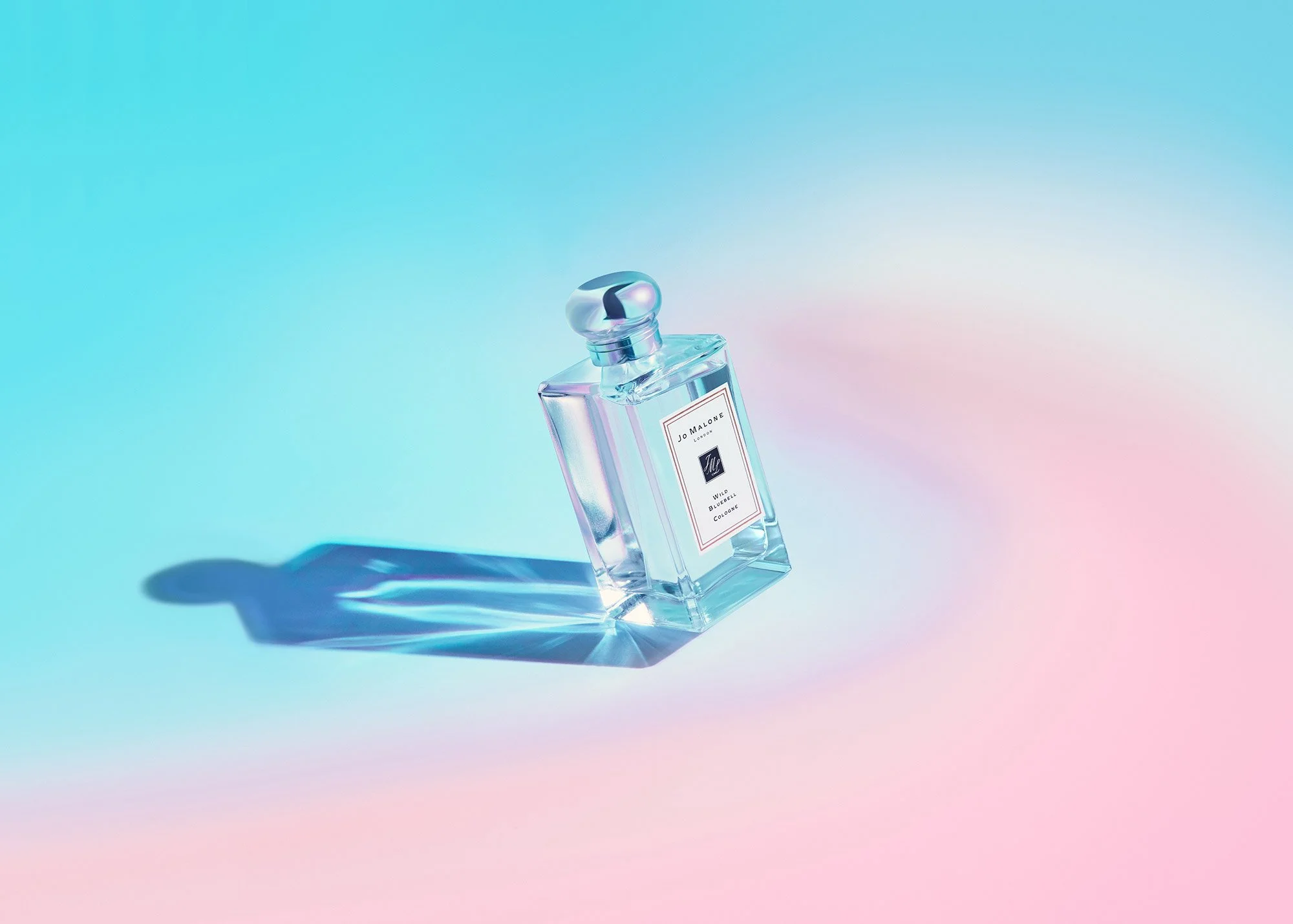 A perfume bottle with a silver cap and a white label, casting colorful shadows on a gradient background of blue, pink, and purple.