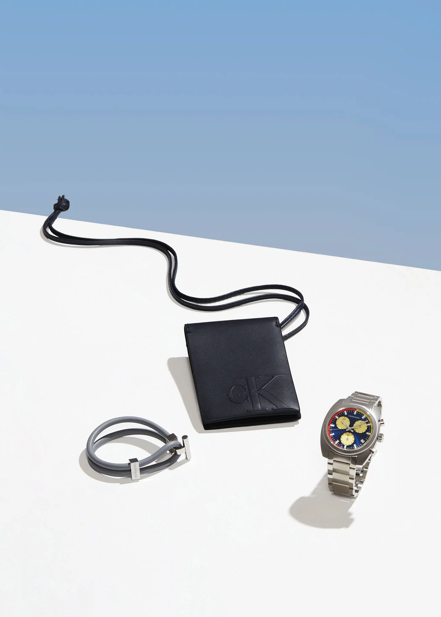 Various fashion accessories including a black leather wallet, a gray bracelet, and a metal watch with a colorful dial on a white surface with a blue background.