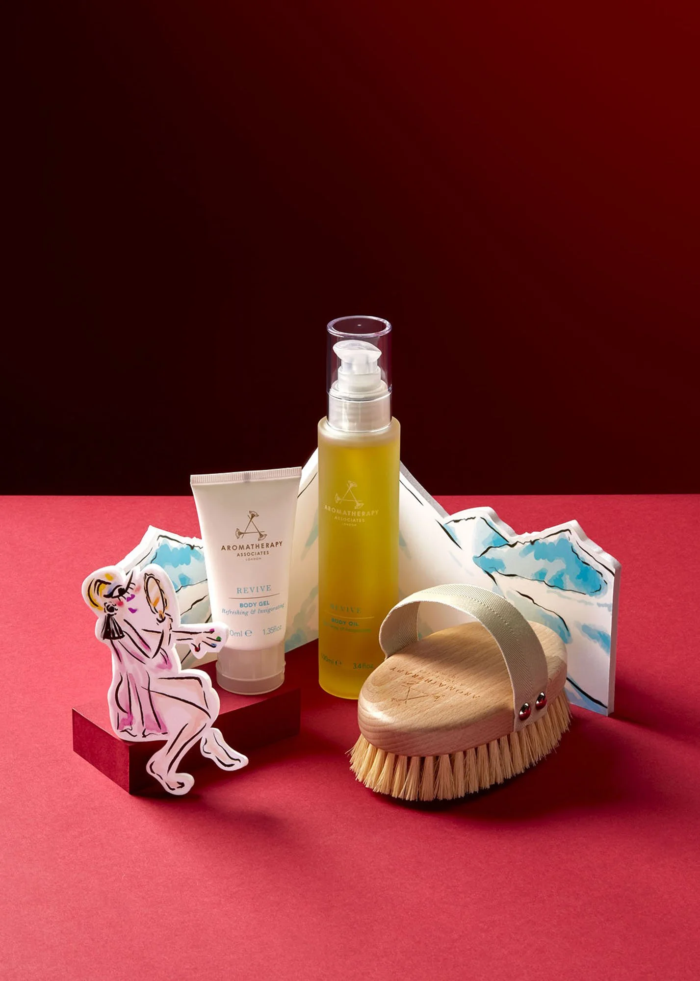 A collection of skincare and body care products including body gel and body oil, a wooden brush with a strap, a decorative paper cutout of a woman in a dress, and a mountain landscape scene on paper, all arranged on a red surface with a dark red back