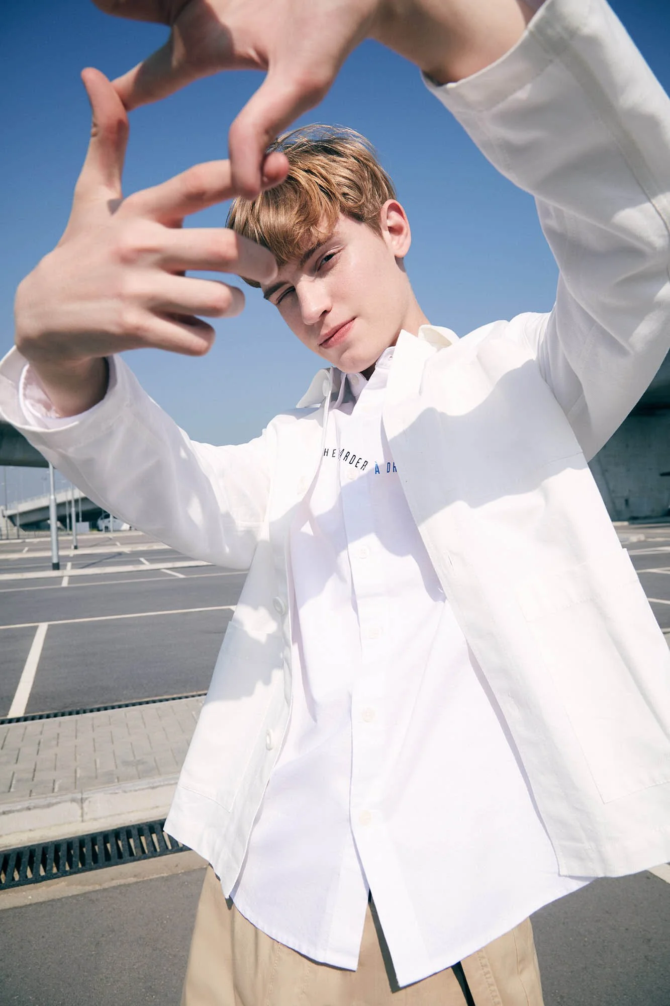 Young man in white shirt and beige pants standing outdoors in a parking lot, making a framing gesture with his hands, with a clear blue sky in the background.