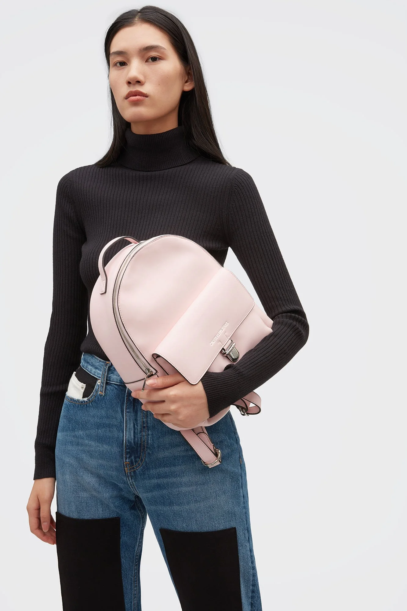 Woman wearing a black turtleneck sweater and blue jeans, holding a pink backpack against a plain white background.