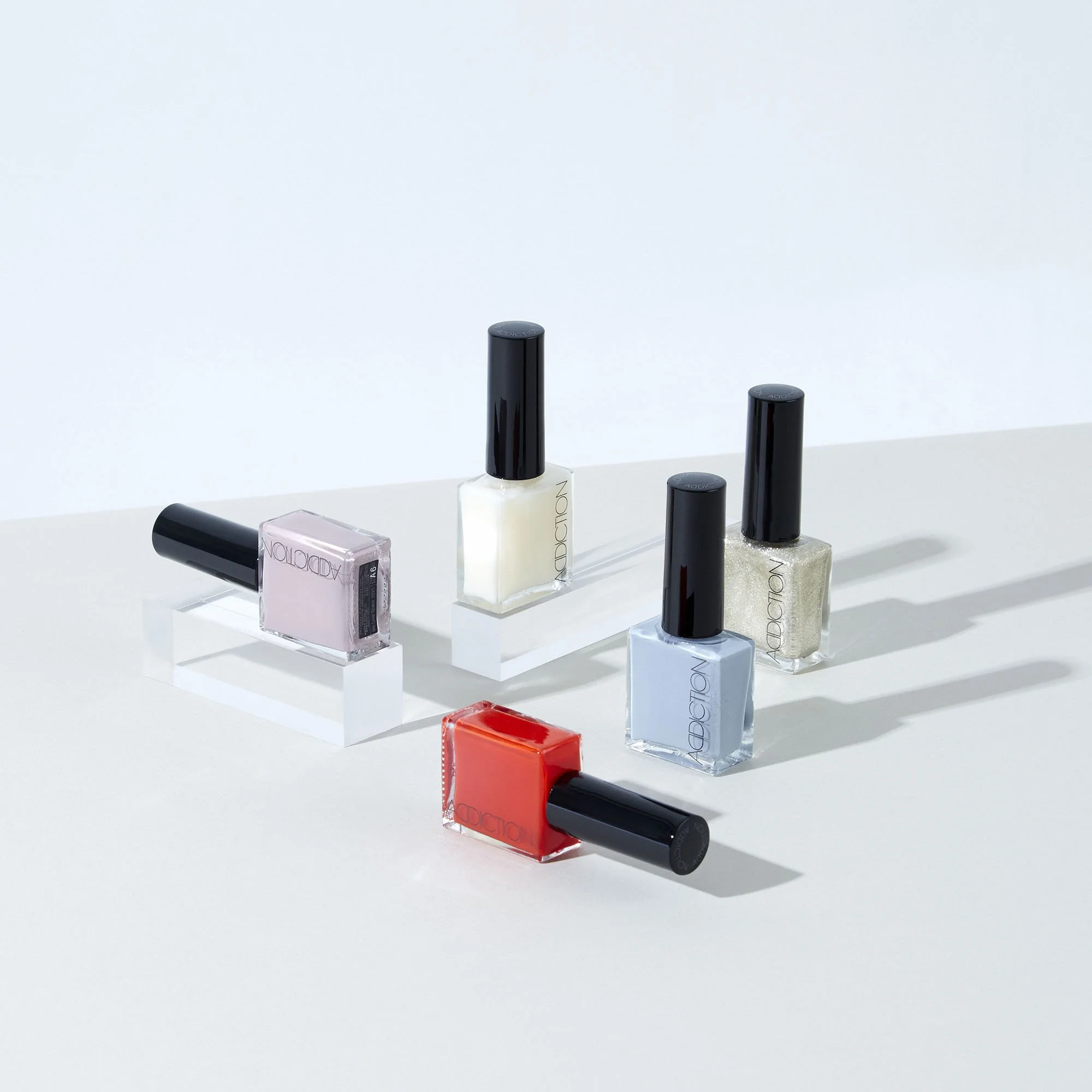 Five bottles of nail polish in various colors arranged on clear acrylic stands on a white surface.