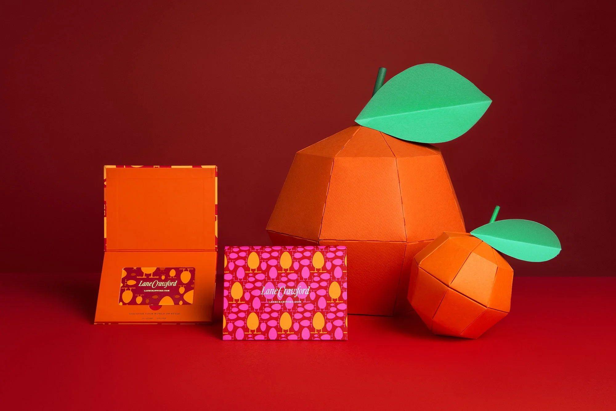 Colorful paper fruit sculptures, including an orange with a green leaf, and smaller orange fruit, along with orange branded boxes, all against a red background.