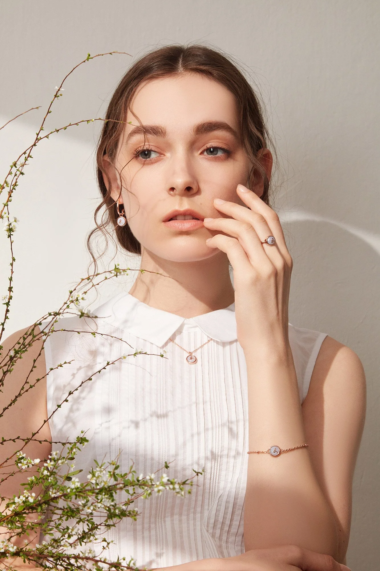 A young woman with wavy brown hair and light skin wearing pearl jewelry and a white sleeveless blouse, standing against a plain background with some greenery in the foreground.