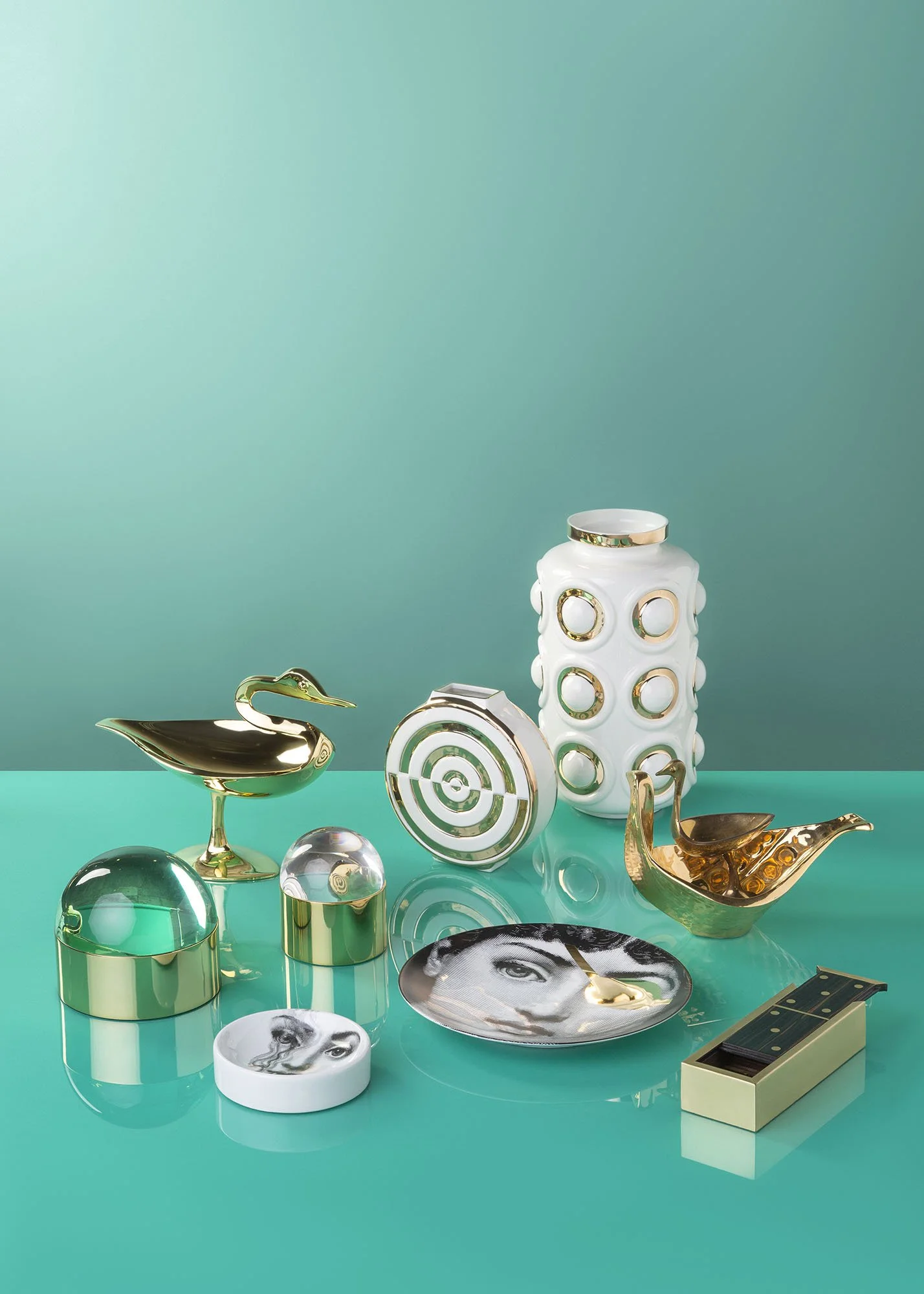 Collection of modern decorative objects including gold and whiteware, a white vase with circular cutouts, a gold swan-shaped container, a white circular container with a face design, a gold dome with a face photo, a compass-like object, and a small g