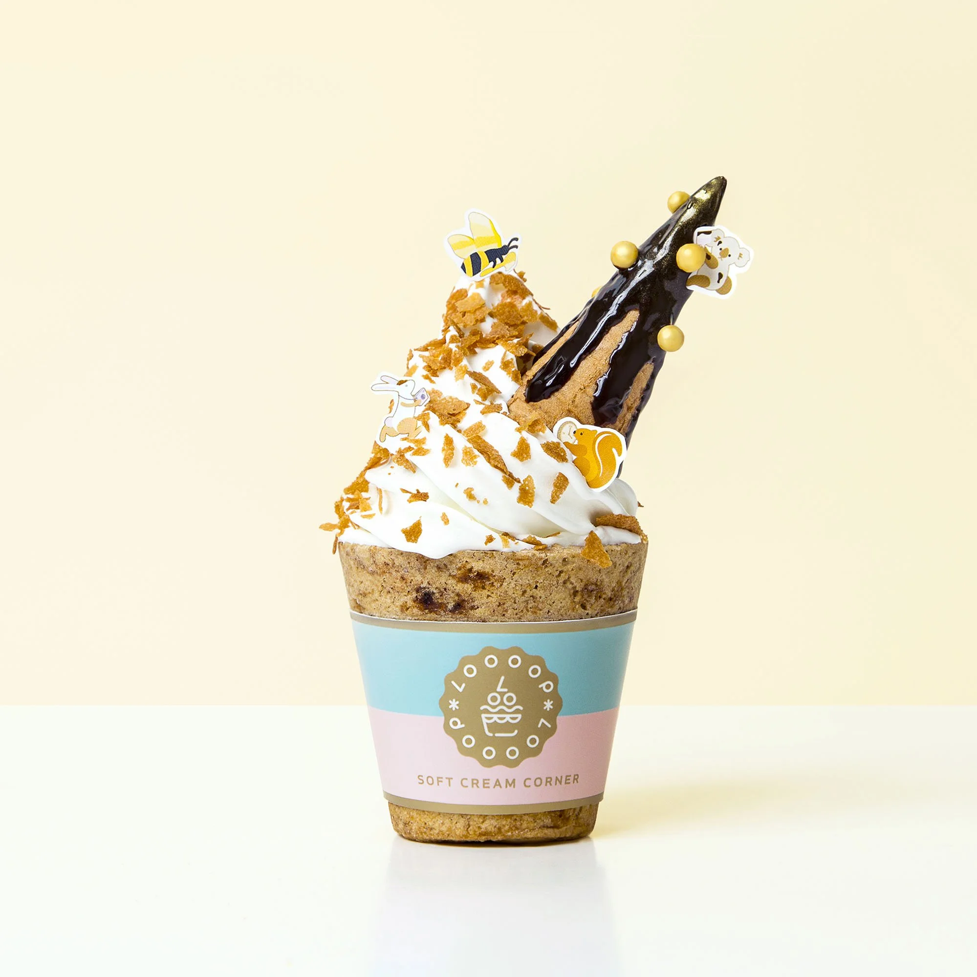 Ice cream sundae in a paper cup with whipped cream, orange sprinkles, a chocolate stick, and small decorative honey bee and squirrel stickers, against a light background.