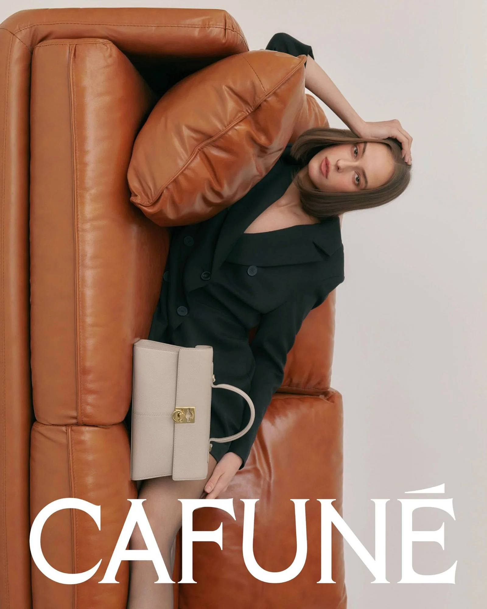 A woman with shoulder-length hair lying on a brown leather couch, wearing a black outfit, with a beige handbag placed beside her, against a plain white wall. The word 'CAFEINE' is overlaid at the bottom of the image.