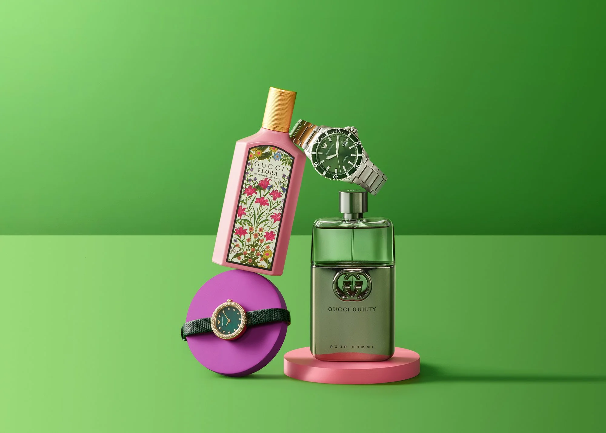 Assortment of luxury perfumes and wristwatches on colorful geometric stands against a green background.