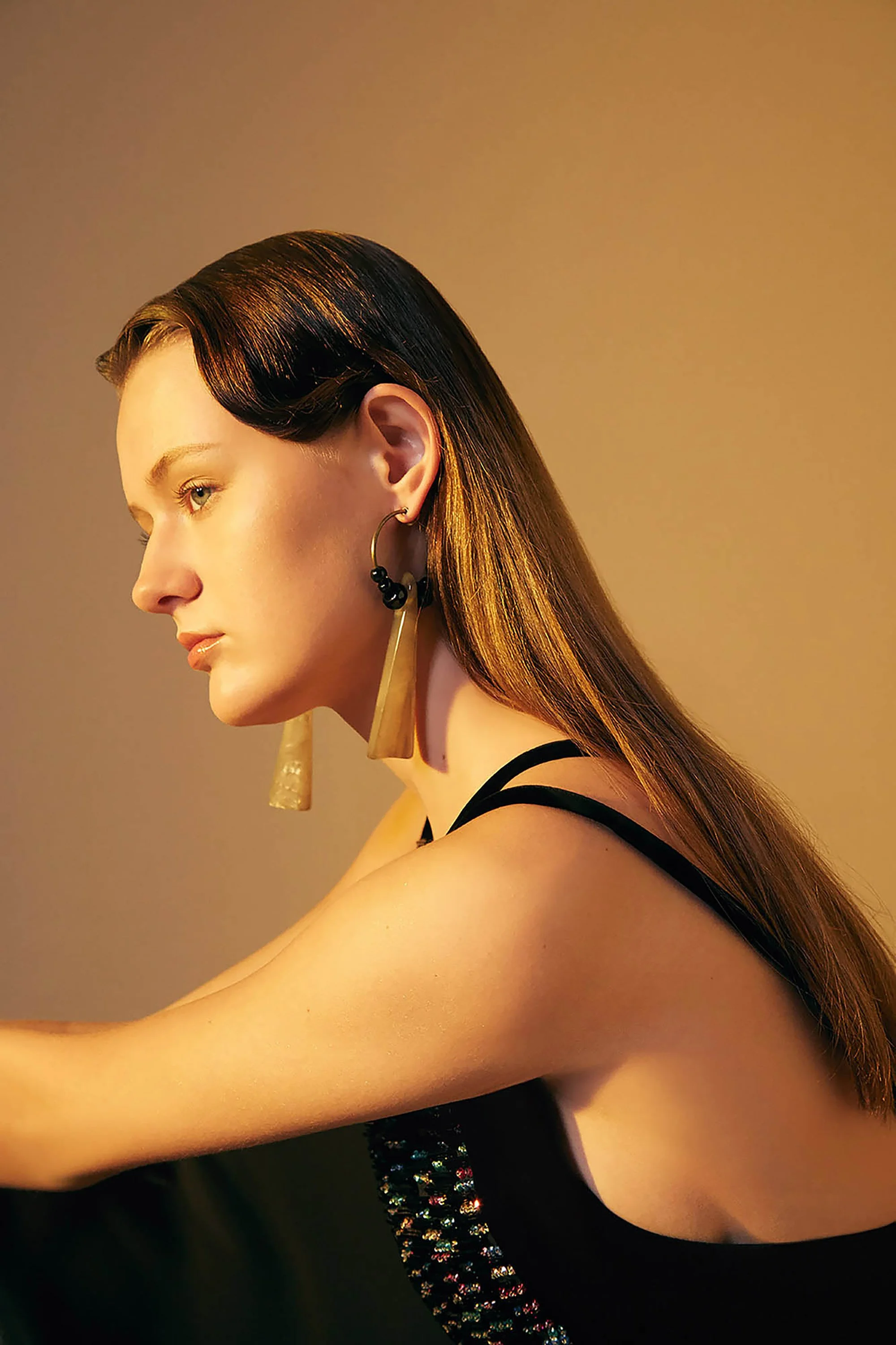 A woman with long brown hair and gold earrings, wearing a black top with thin straps, is looking to her left against a plain wall with warm lighting.