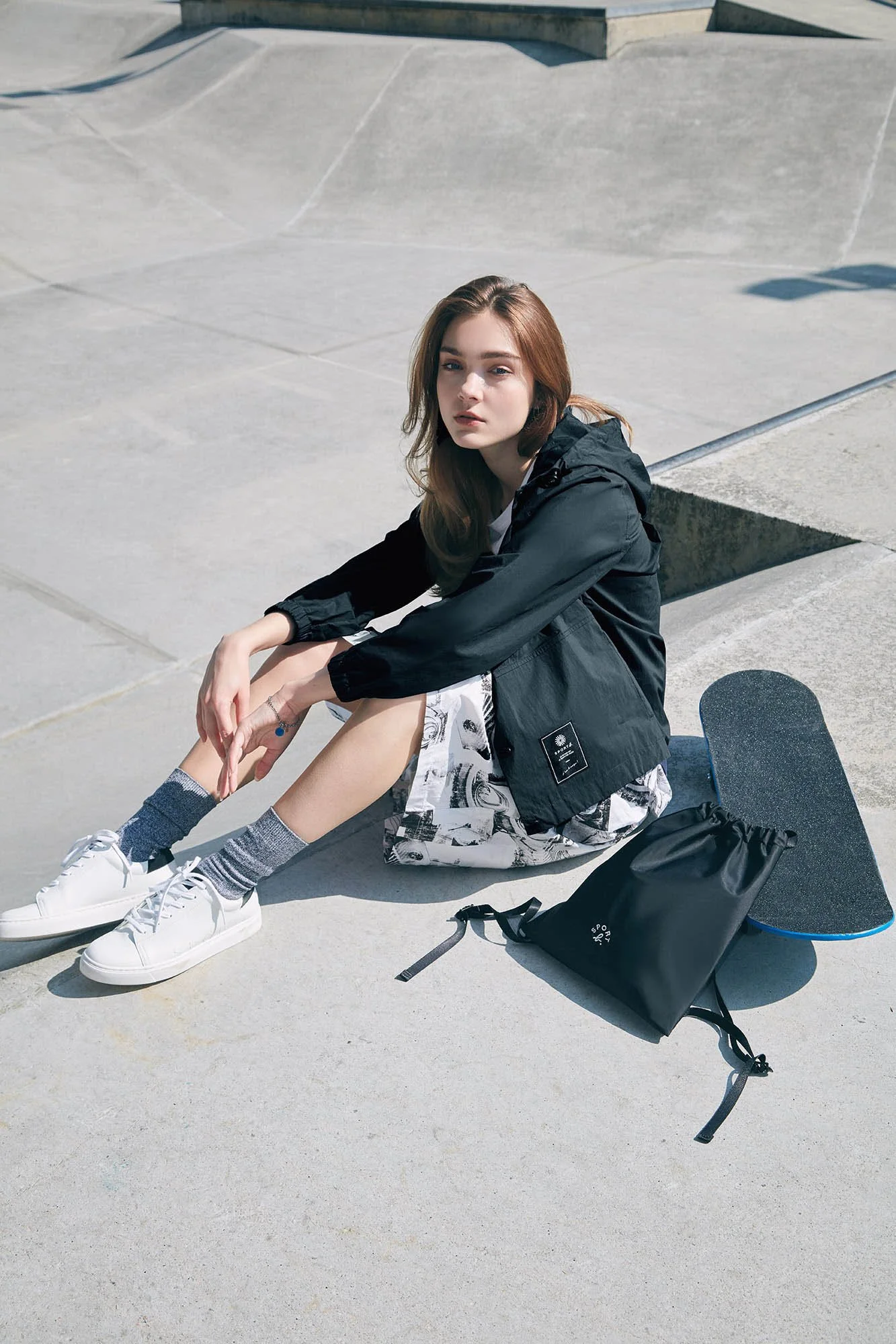 A young woman with light skin and brown hair sits on the ground at a skate park, next to her skateboard, wearing a black jacket, printed shorts, and white sneakers, with a black drawstring bag nearby.
