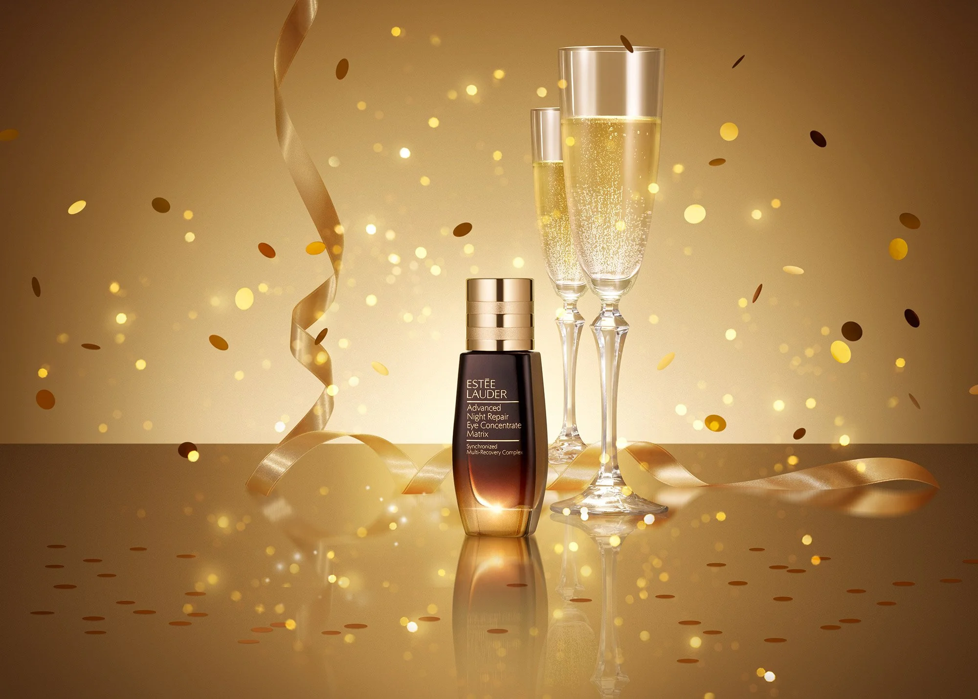 A bottle of Estée Lauder Advanced Night Repair Eye Concentrate Matrix surrounded by two champagne glasses and gold party streamers and confetti on a reflective gold surface.