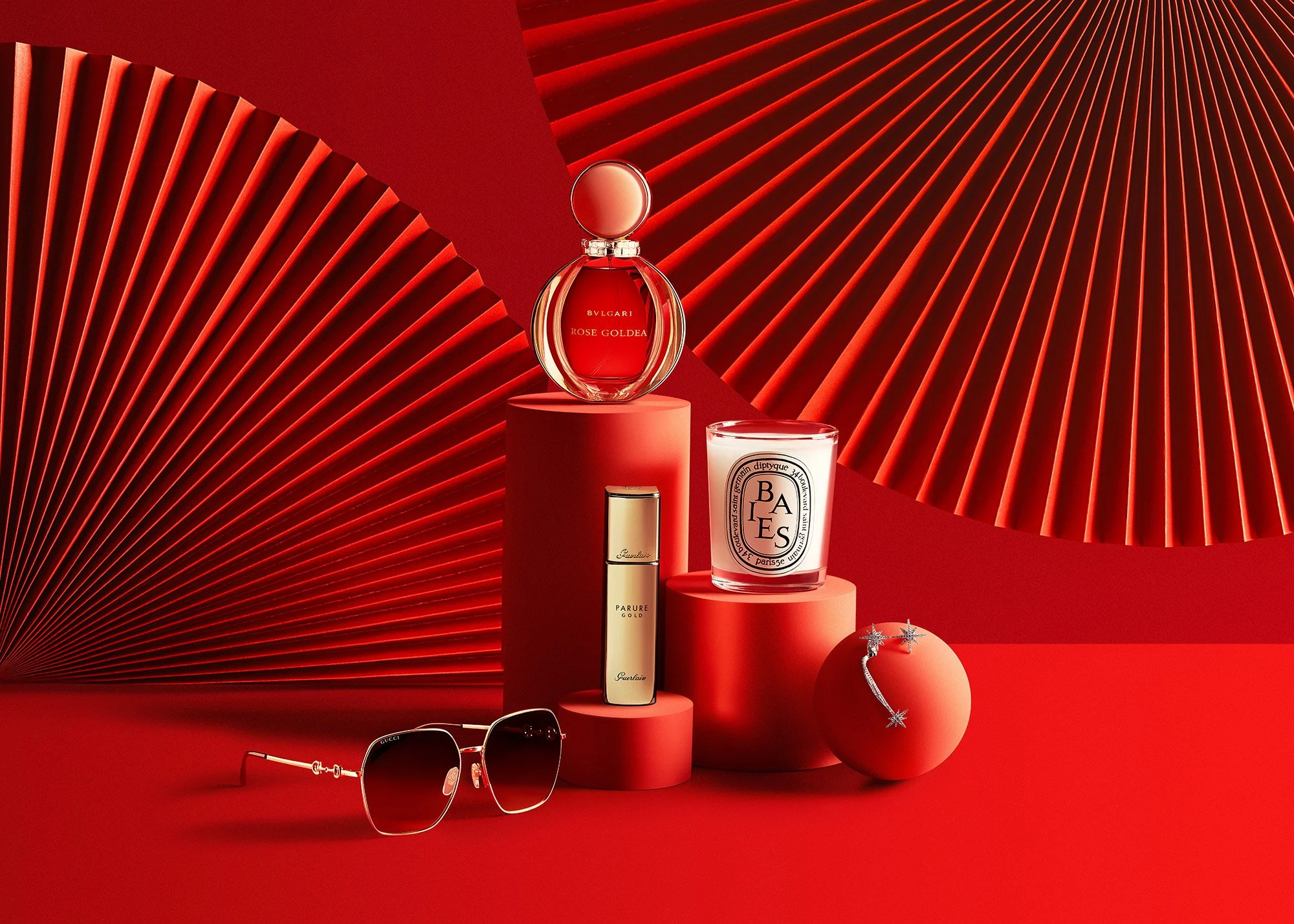 A collection of luxury products on red pedestals, including perfume bottles, a candle, sunglasses, and jewelry, set against a red background with fan-shaped geometric designs.
