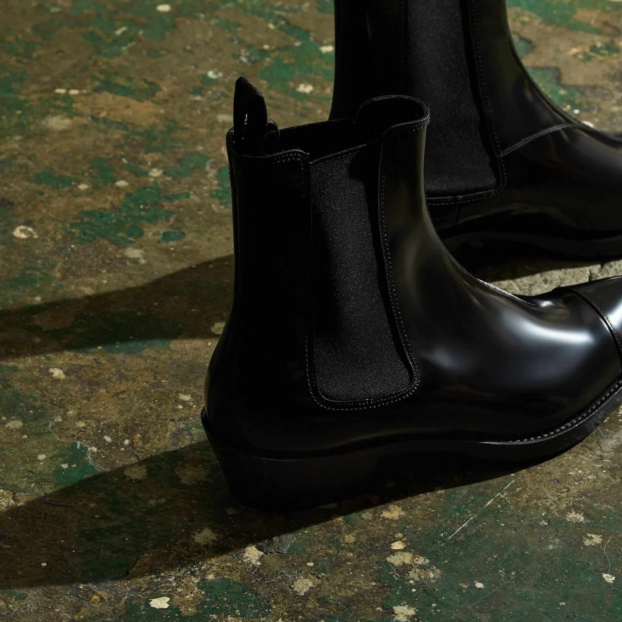 Close-up of a pair of shiny black leather Chelsea boots on a worn, textured floor with green and beige paint splatters.