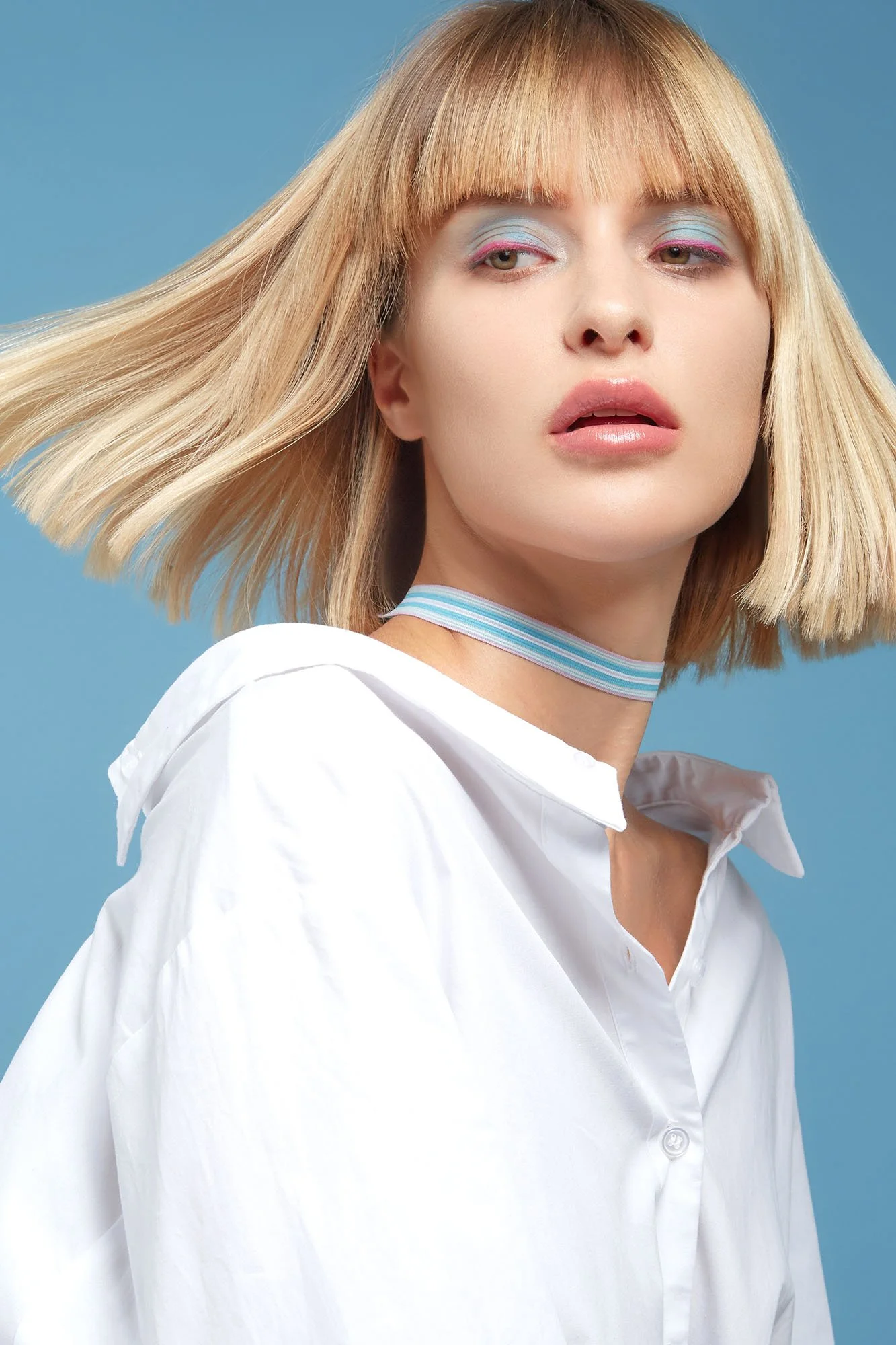 A young woman with blonde hair, wearing pastel eyeshadow, a striped choker, and a white shirt, against a blue background.
