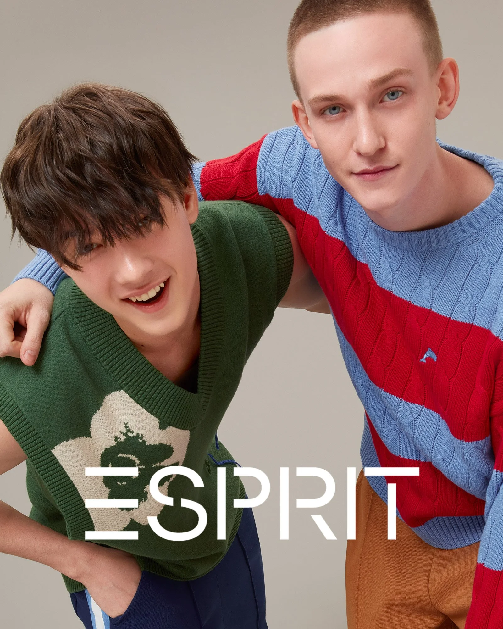 Two young men, one with brown hair and the other with blonde hair, smiling and leaning into each other. The brown-haired man is wearing a green sleeveless sweater with a white and green graphic, and the blonde-haired man is wearing a blue and red swe