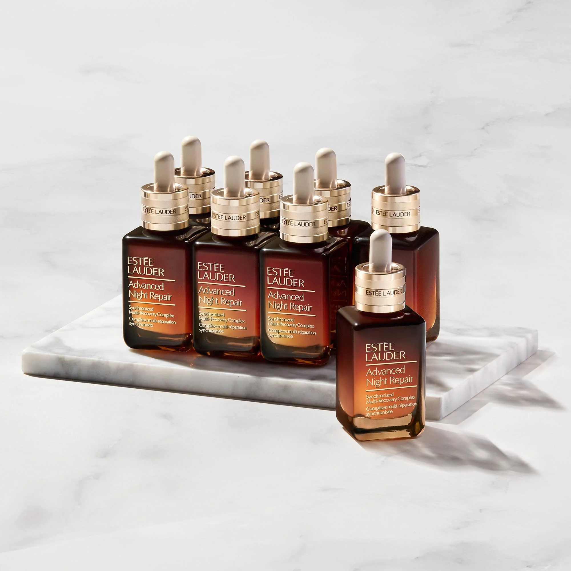 Six bottles of Estée Lauder Advanced Night Repair serum arranged on a white marble slab against a white background.