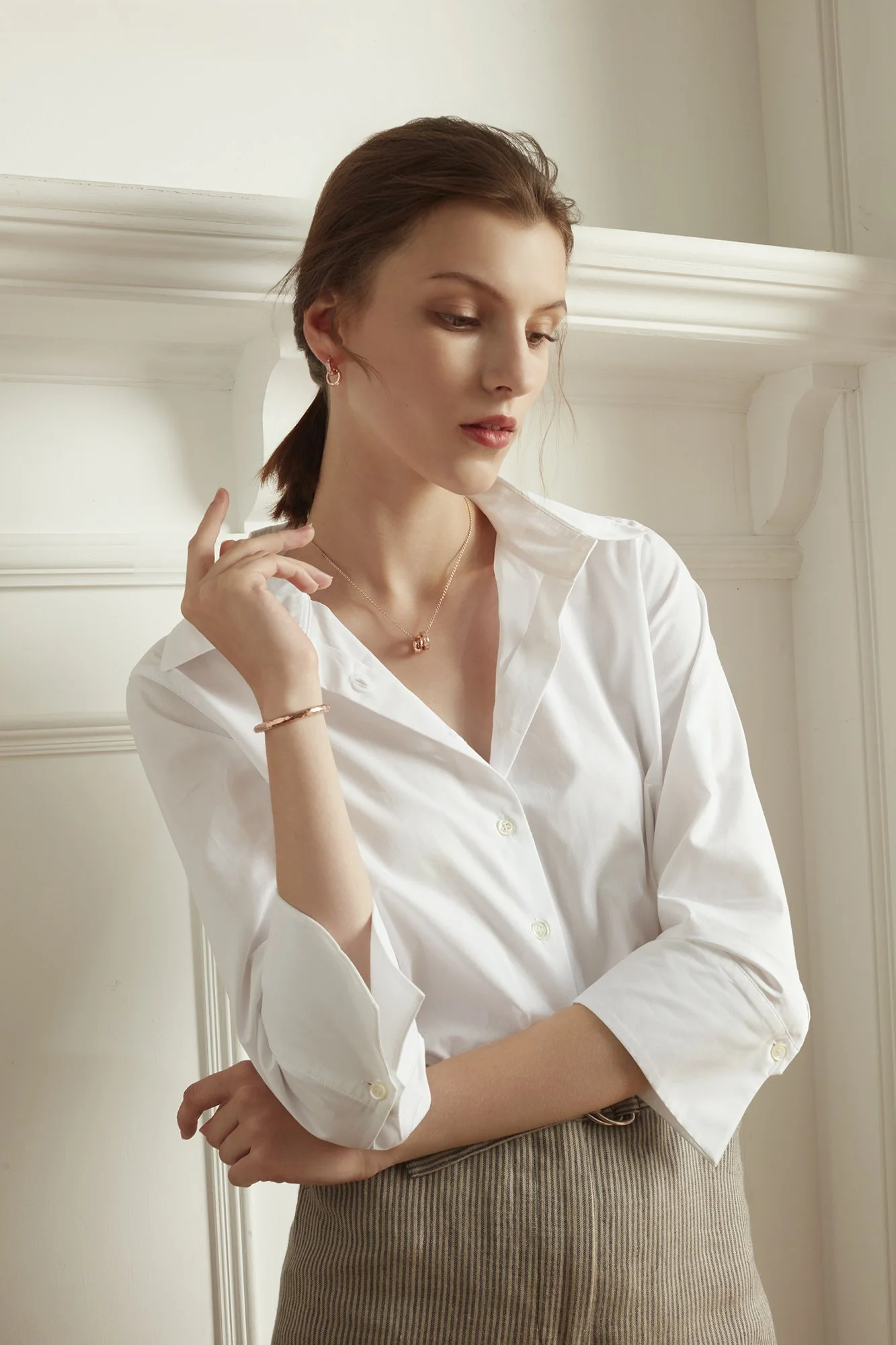A young woman with brown hair in a ponytail, wearing a white button-up shirt and beige skirt, standing indoors with a neutral background, looking down thoughtfully.