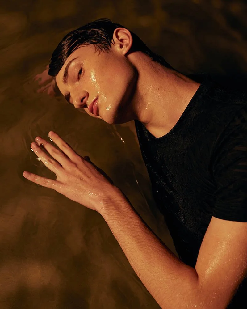 A young man with wet hair and face lying on his side in water, wearing a black shirt, with one hand touching the water's surface.
